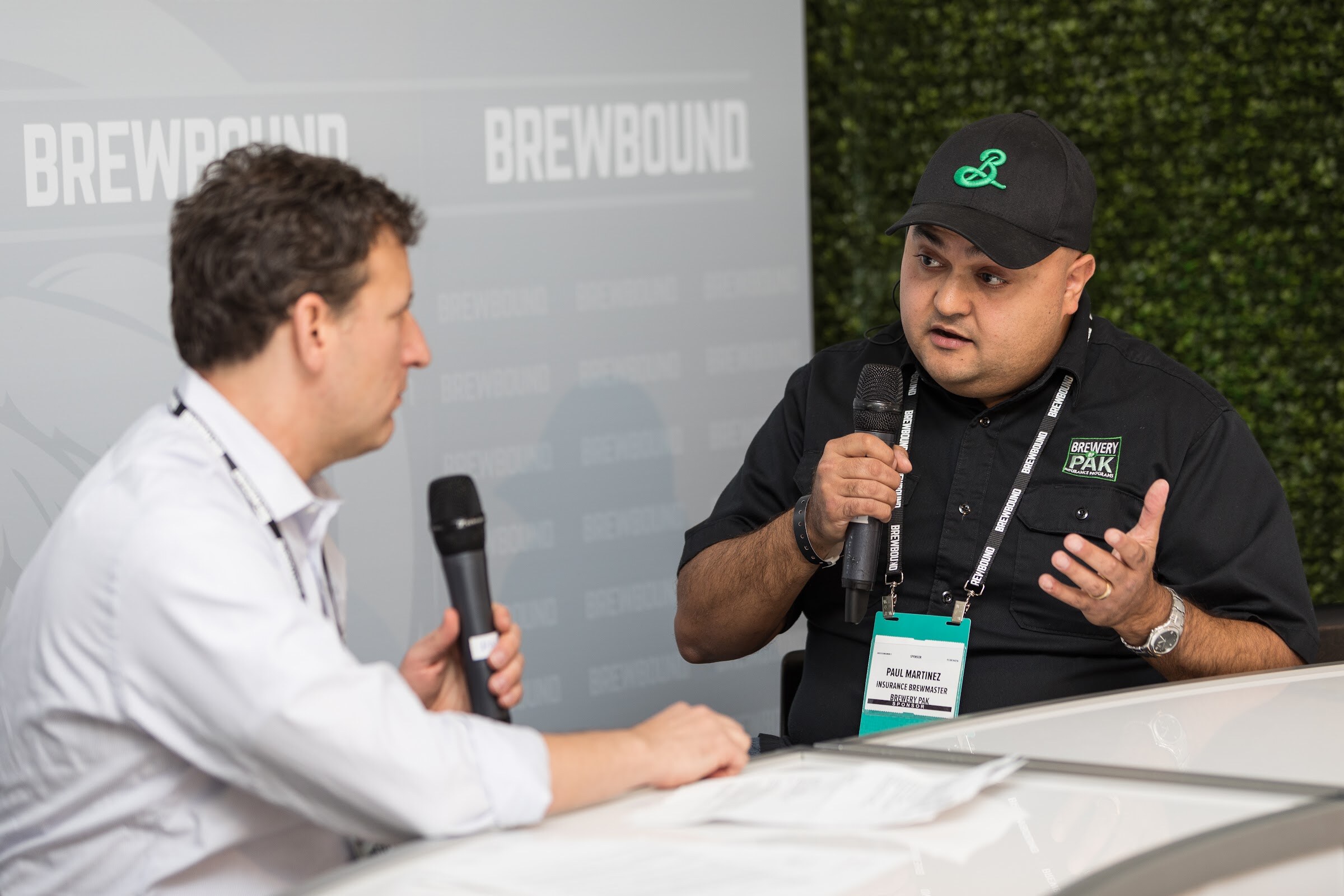 A man in a black cap and black Brewery Pak shirt holds a microphone and gestures while a man in a white shirt holds a microphone beside him.