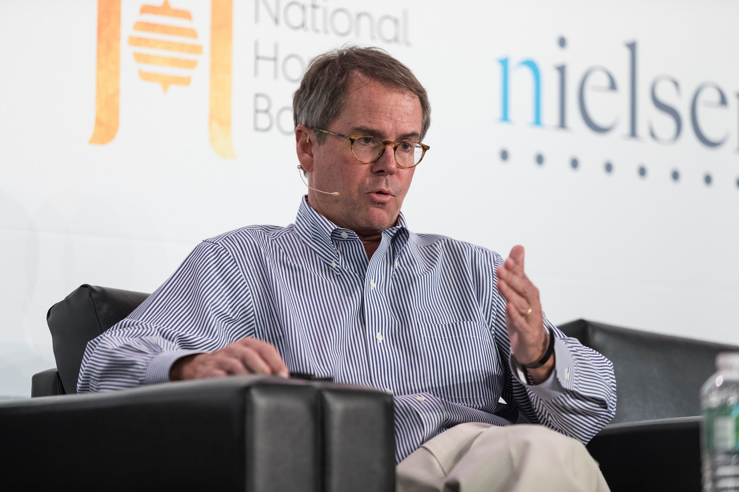 A man in glasses and a blue striped shirt gestures with one hand from a black armchair in front of National Honey Board and Nielsen signs.