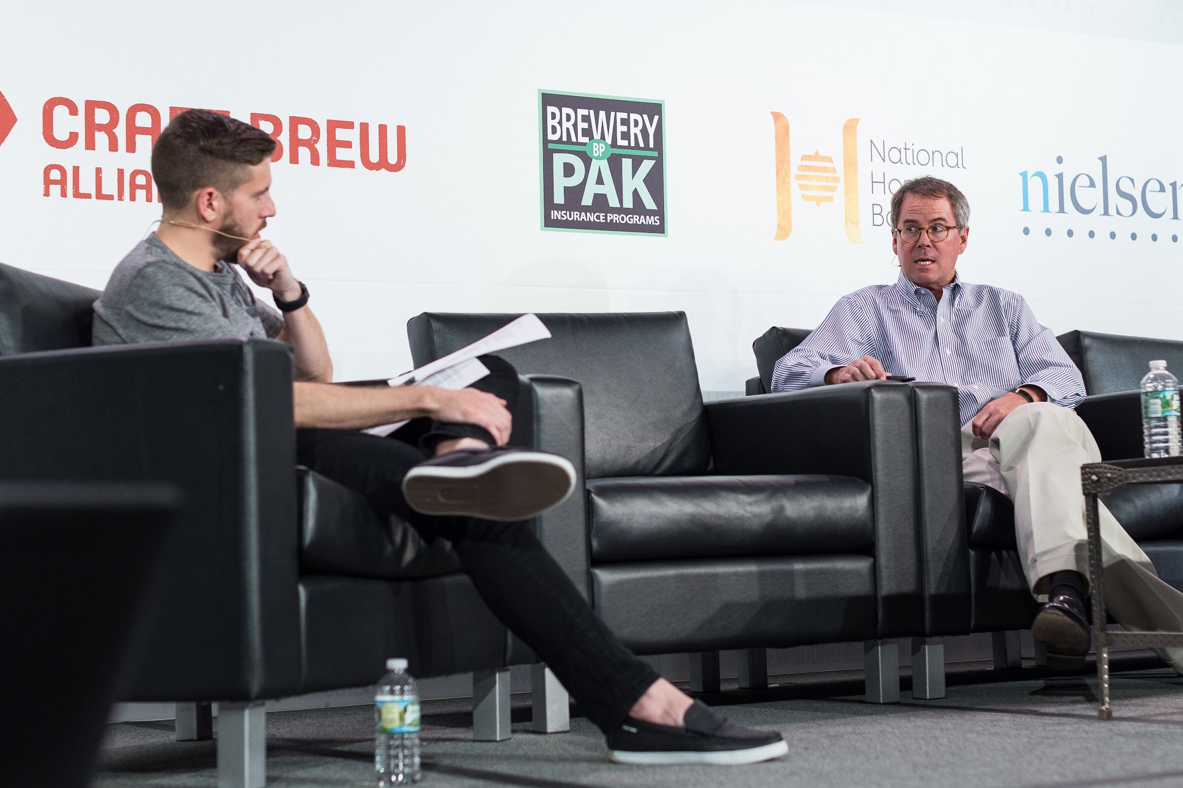 A host in a gray shirt holding papers and a man in a striped shirt sit in black armchairs on stage in front of Craft Brew Alliance, Brewery Pak and Nielsen signs.