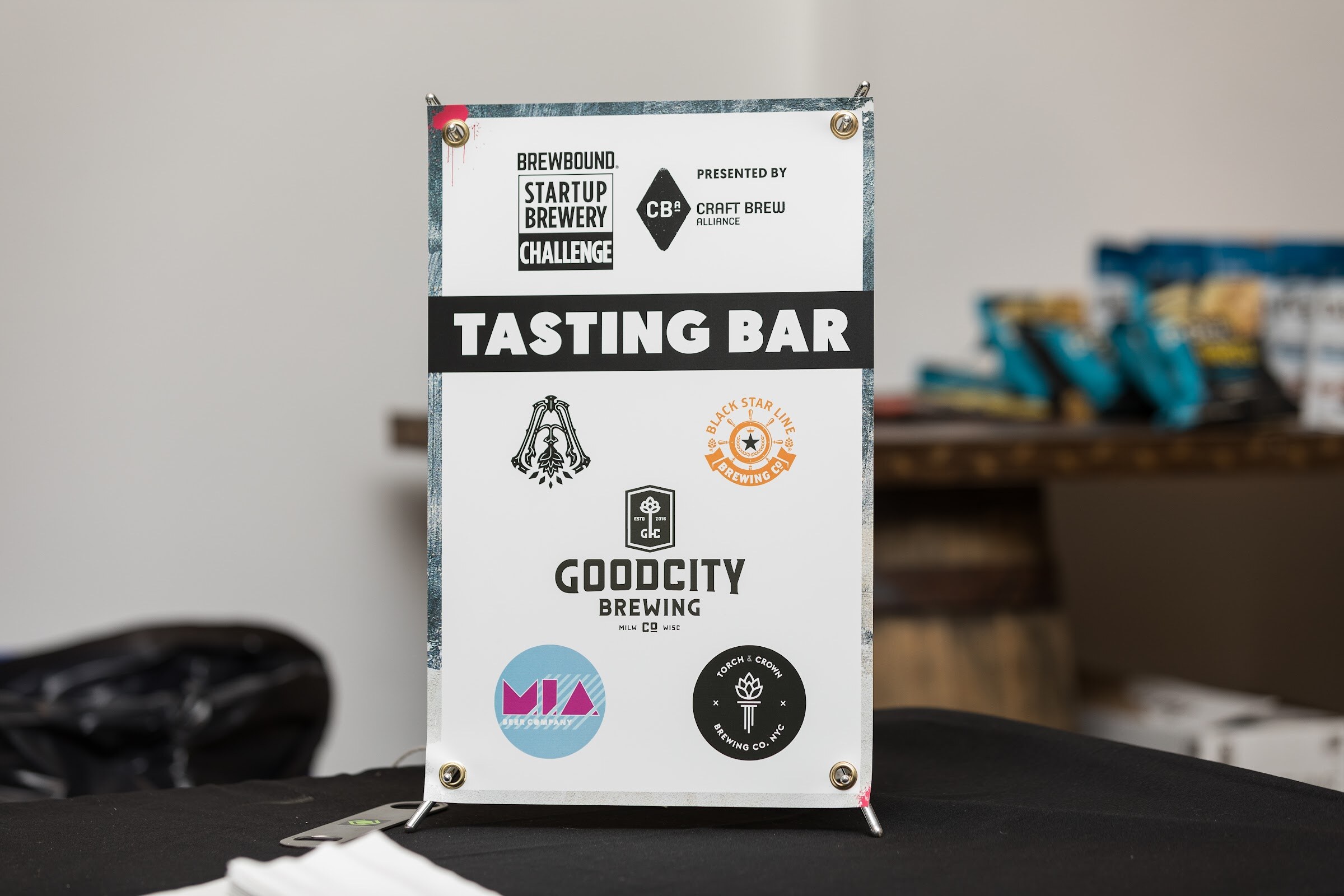 A standing sign reading Tasting Bar with the Brewbound Startup Brewery Challenge logo, Presented by Craft Brew Alliance, and logos for Armada, Black Star Line, Good City, M.I.A. and Torch & Crown breweries.