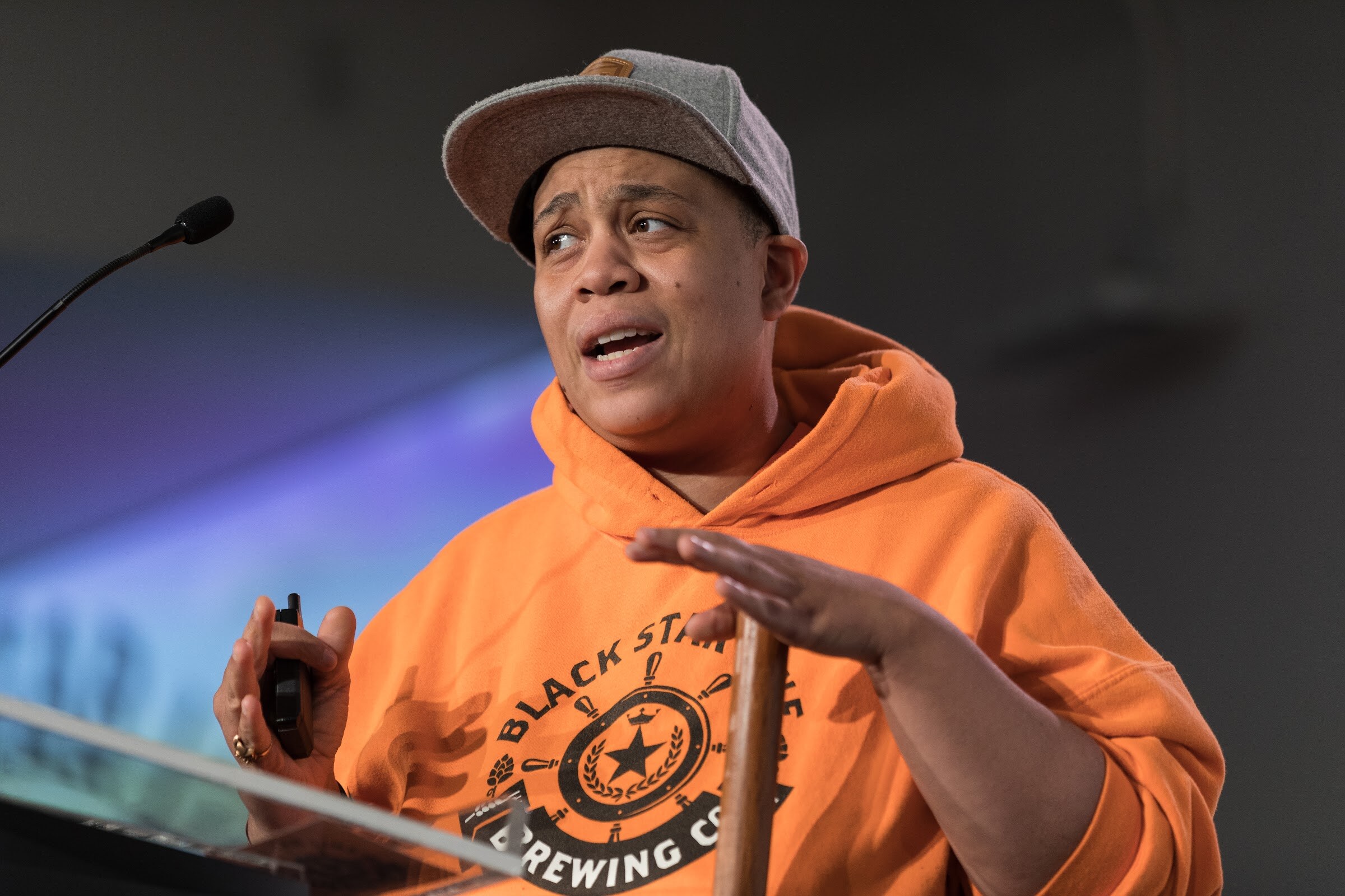 A person in a gray cap and orange Black Star Line Brewing hoodie holds a clicker and speaks at a podium.