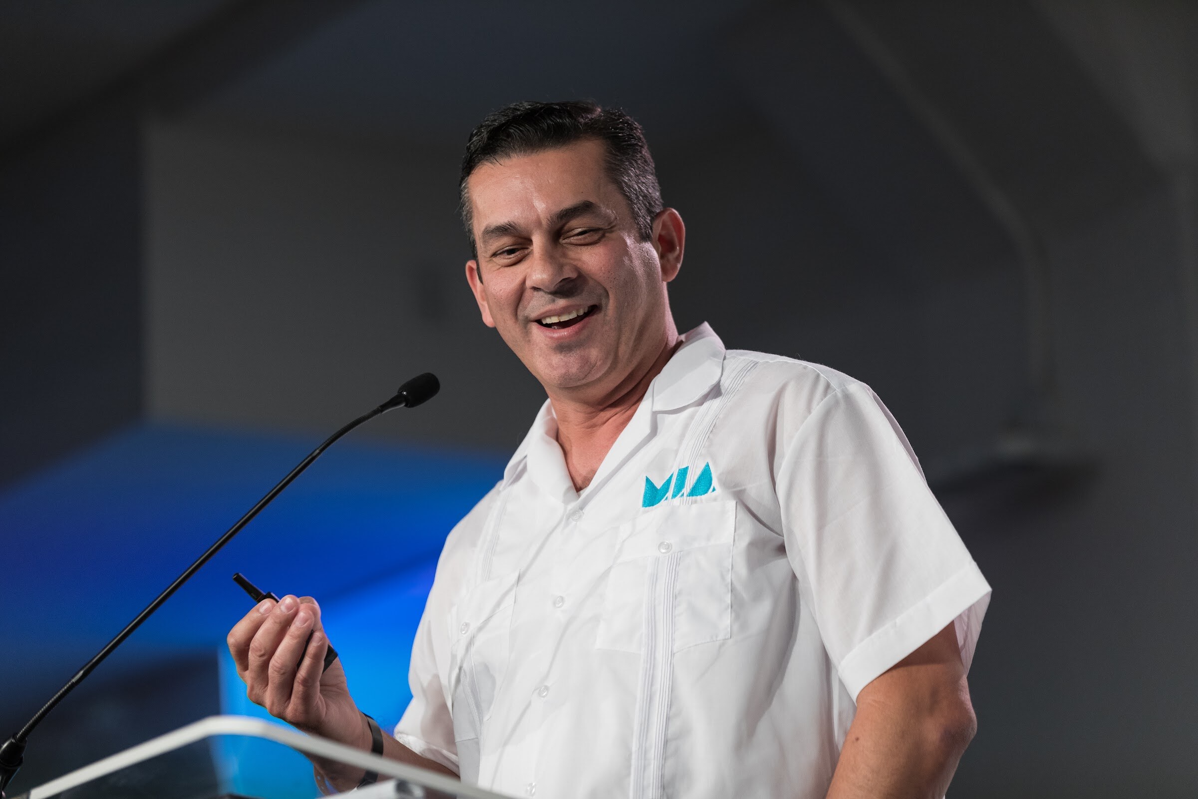 A smiling man in a white short-sleeved guayabera shirt with a teal MIA logo holds a clicker beside a microphone.