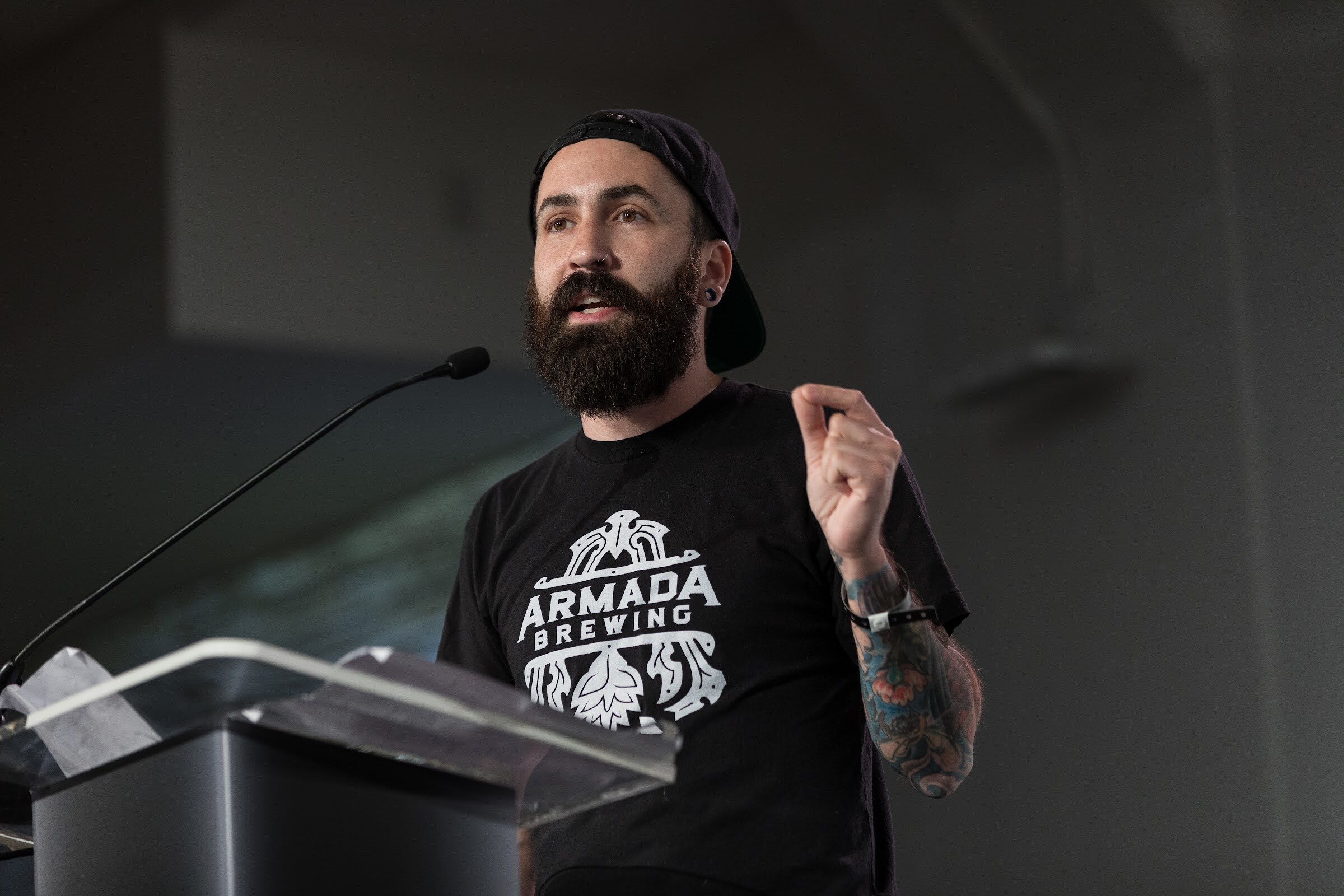 A bearded man in a backward cap and black Armada Brewing T-shirt gestures while speaking at a clear podium.