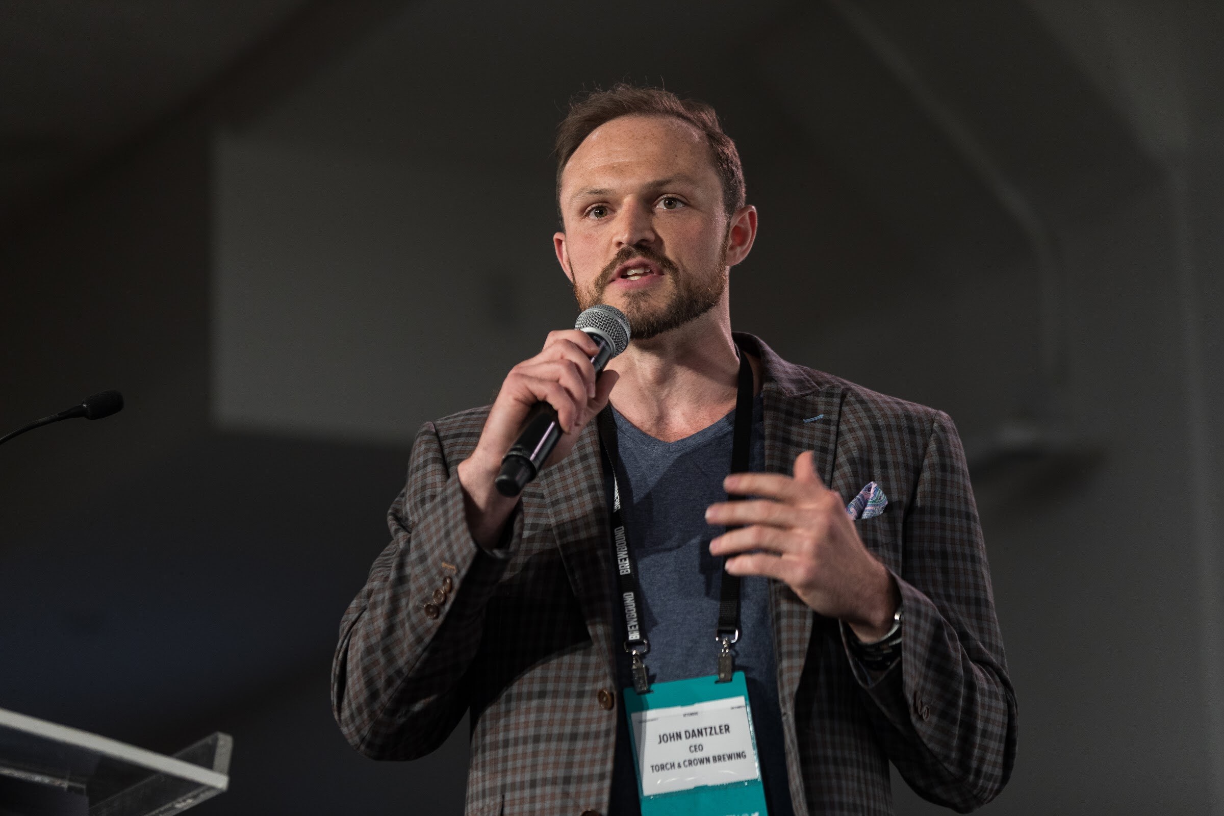 A man in a plaid blazer with a conference badge reading Torch & Crown Brewing holds a microphone and gestures while speaking.