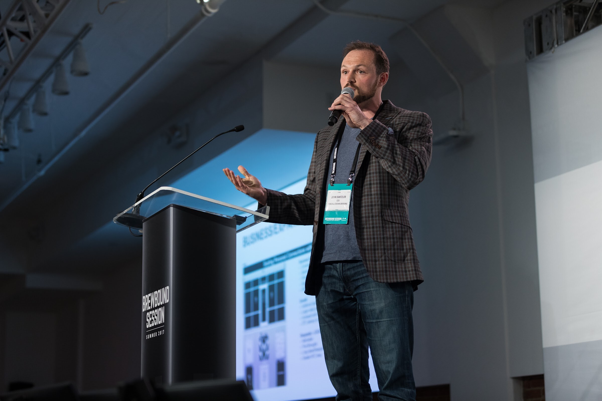 A man in a plaid blazer with a microphone gestures on stage next to a black Brewbound Session Summer 2017 podium with a presentation slide projected behind him.