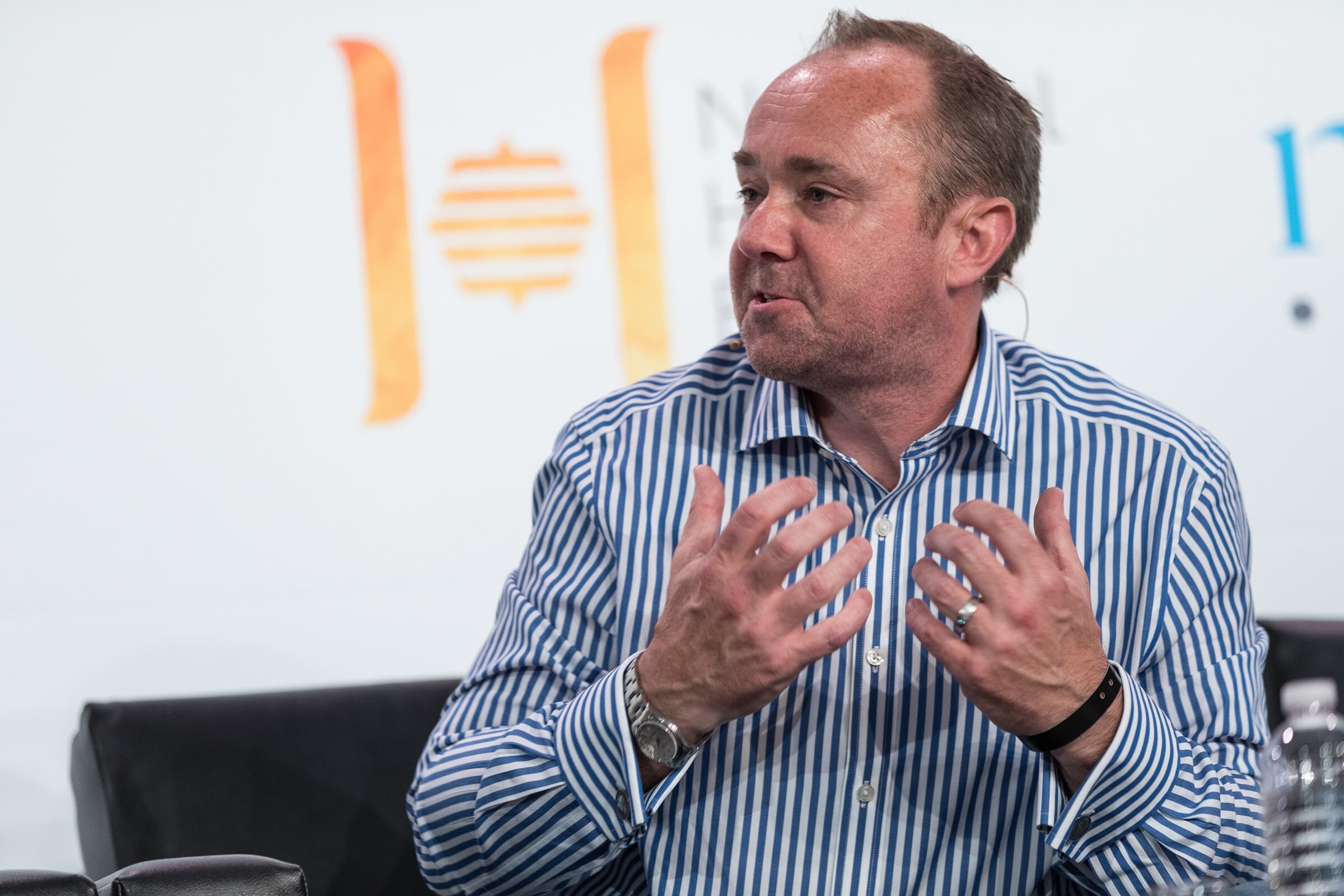 A man in a blue striped shirt gestures with both hands from a black chair in front of a National Honey Board sign.