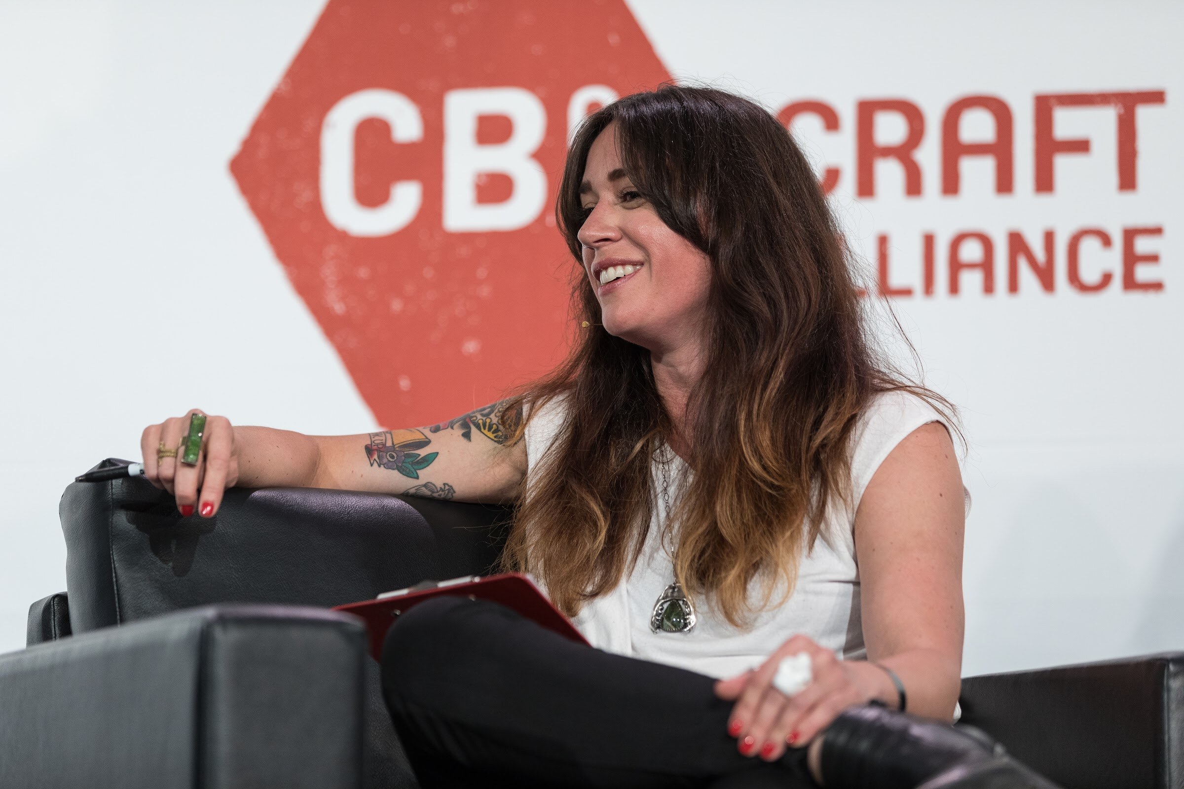 A woman with long hair in a white sleeveless top smiles while seated in a black armchair with a red clipboard in front of a Craft Brew Alliance sign.