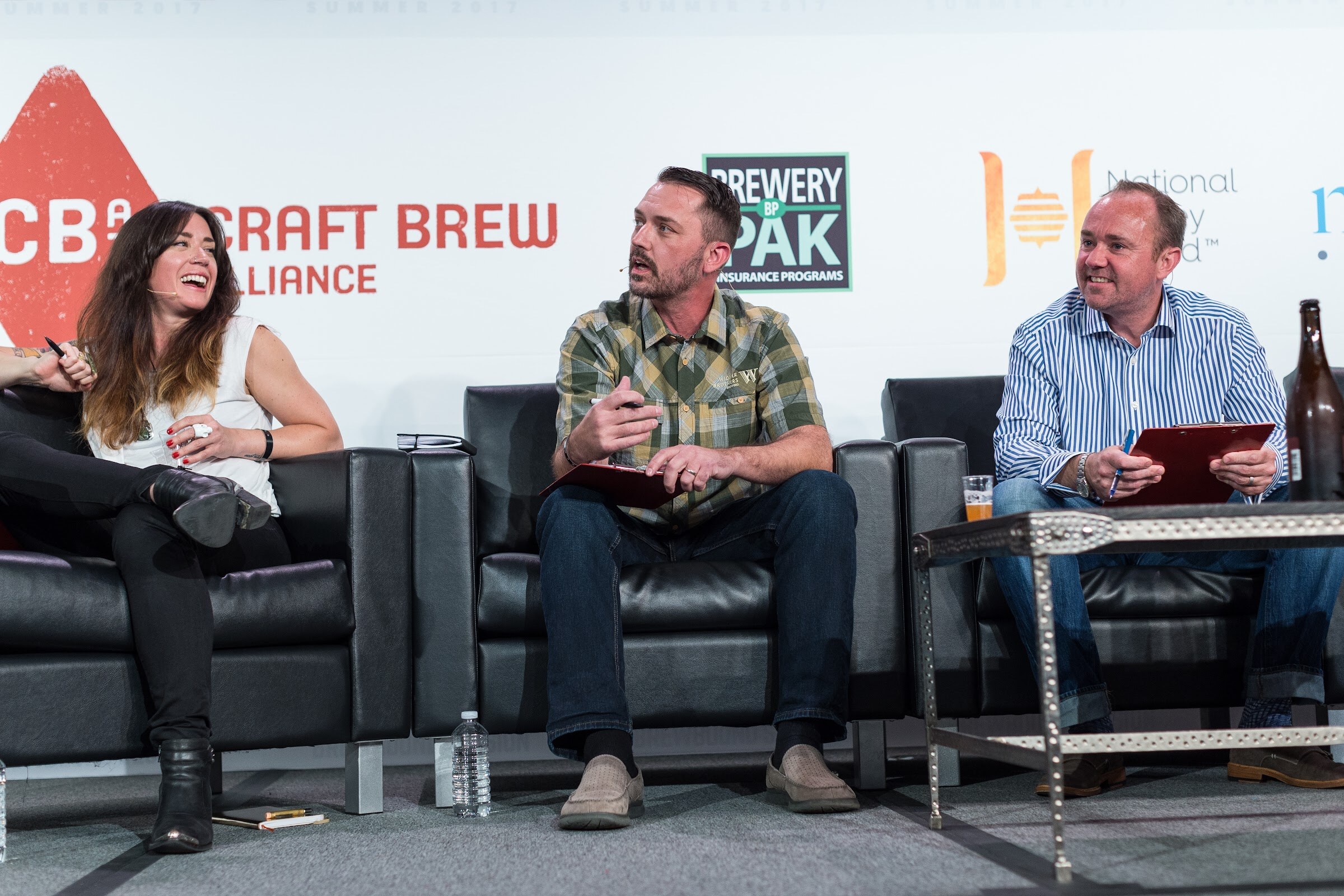 A woman and two men with clipboards sit on black couches in front of Craft Brew Alliance, Brewery Pak and National Honey Board signs; one man in a plaid shirt speaks while the others laugh.