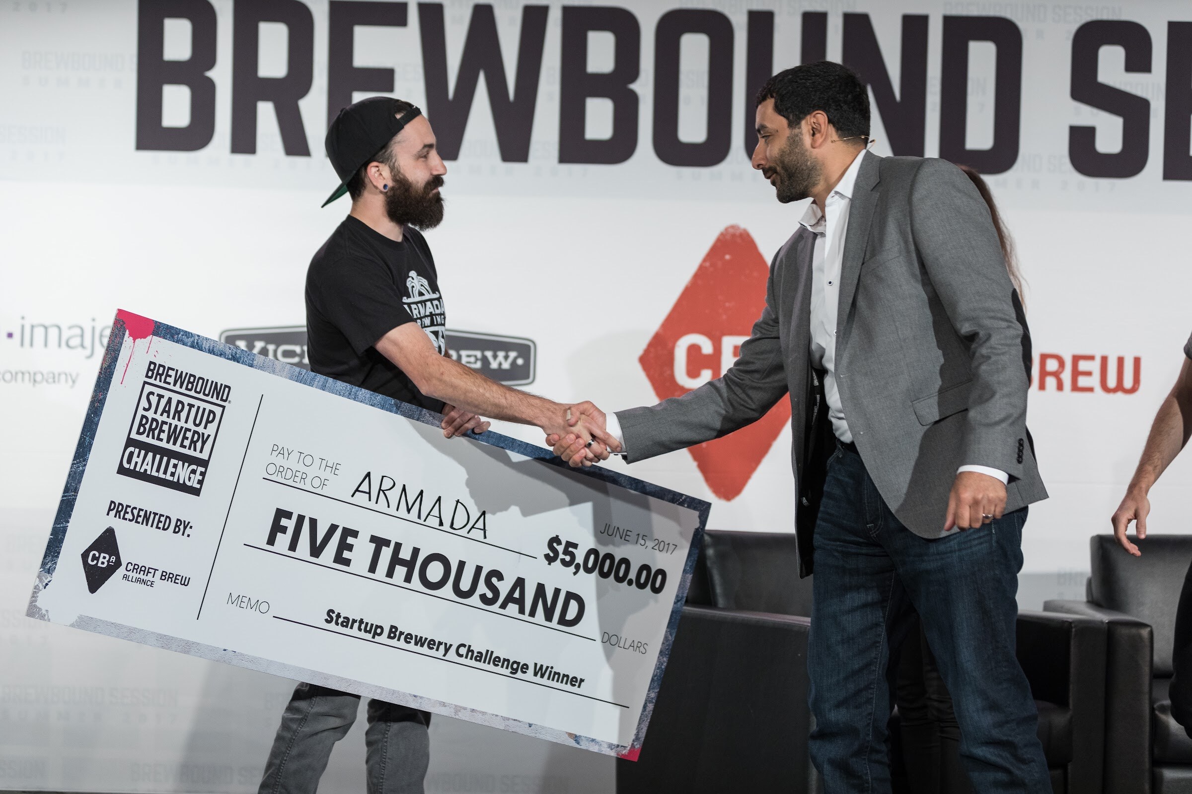 A bearded man holds a large check for five thousand dollars made out to Armada while shaking hands with a man in a gray blazer.