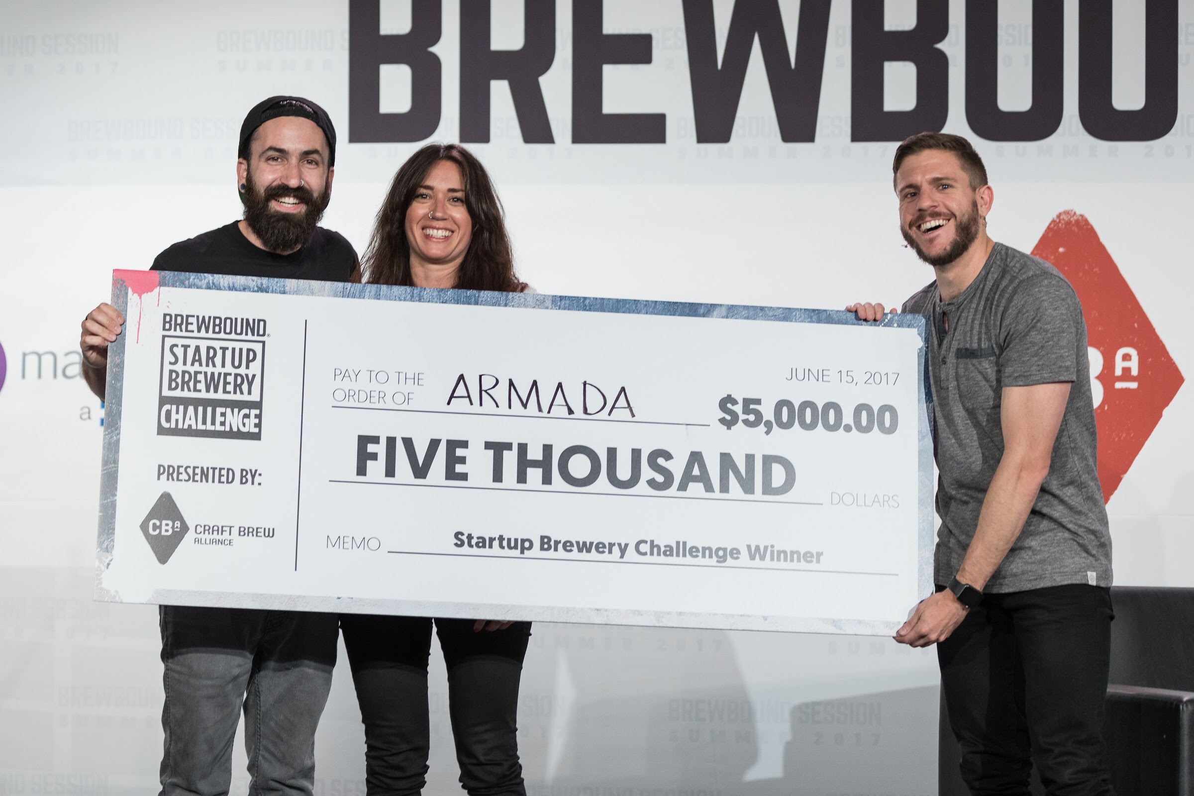 Three smiling people hold an oversized five thousand dollar check made out to Armada for the Startup Brewery Challenge Winner.