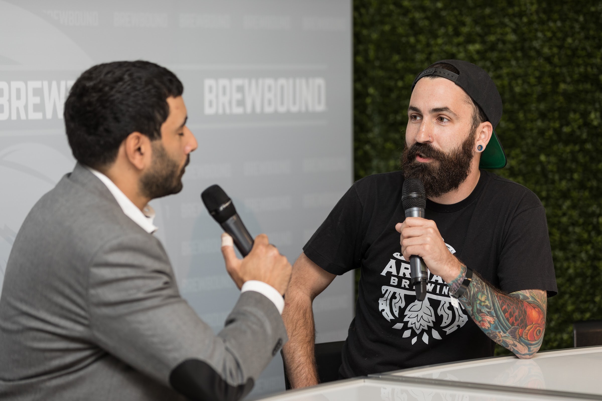 A bearded man in a backward cap and Armada Brewing shirt holds a microphone and talks with a man in a gray blazer at a white desk in front of a hedge wall.