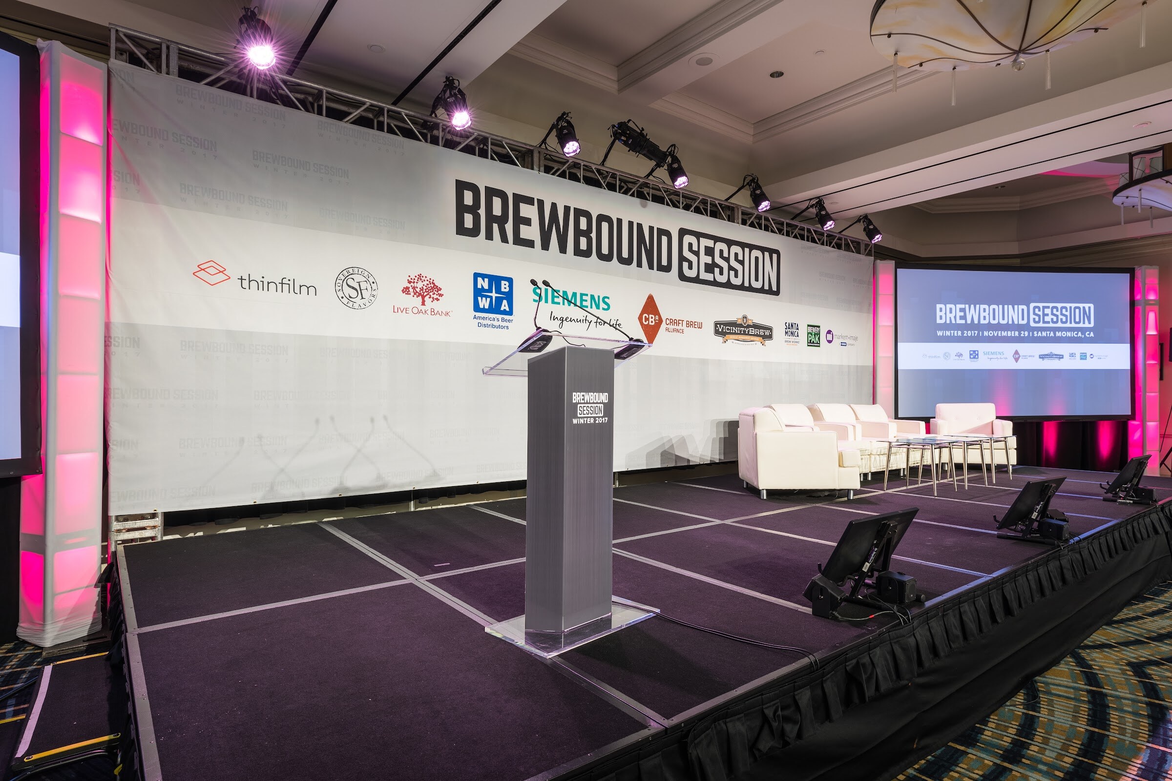 An empty stage with a gray Brewbound Session Winter 2017 podium, a row of white sofas and glass tables, magenta lighting and a Brewbound Session banner with sponsor logos.