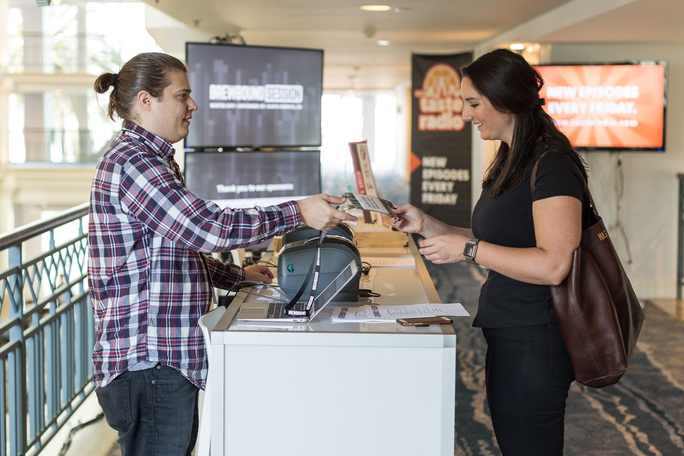 A man in a purple plaid shirt with a bun hands a printed card across a white counter with a label printer and laptop to a smiling woman with a brown shoulder bag, with a Taste Radio banner and screens behind them.