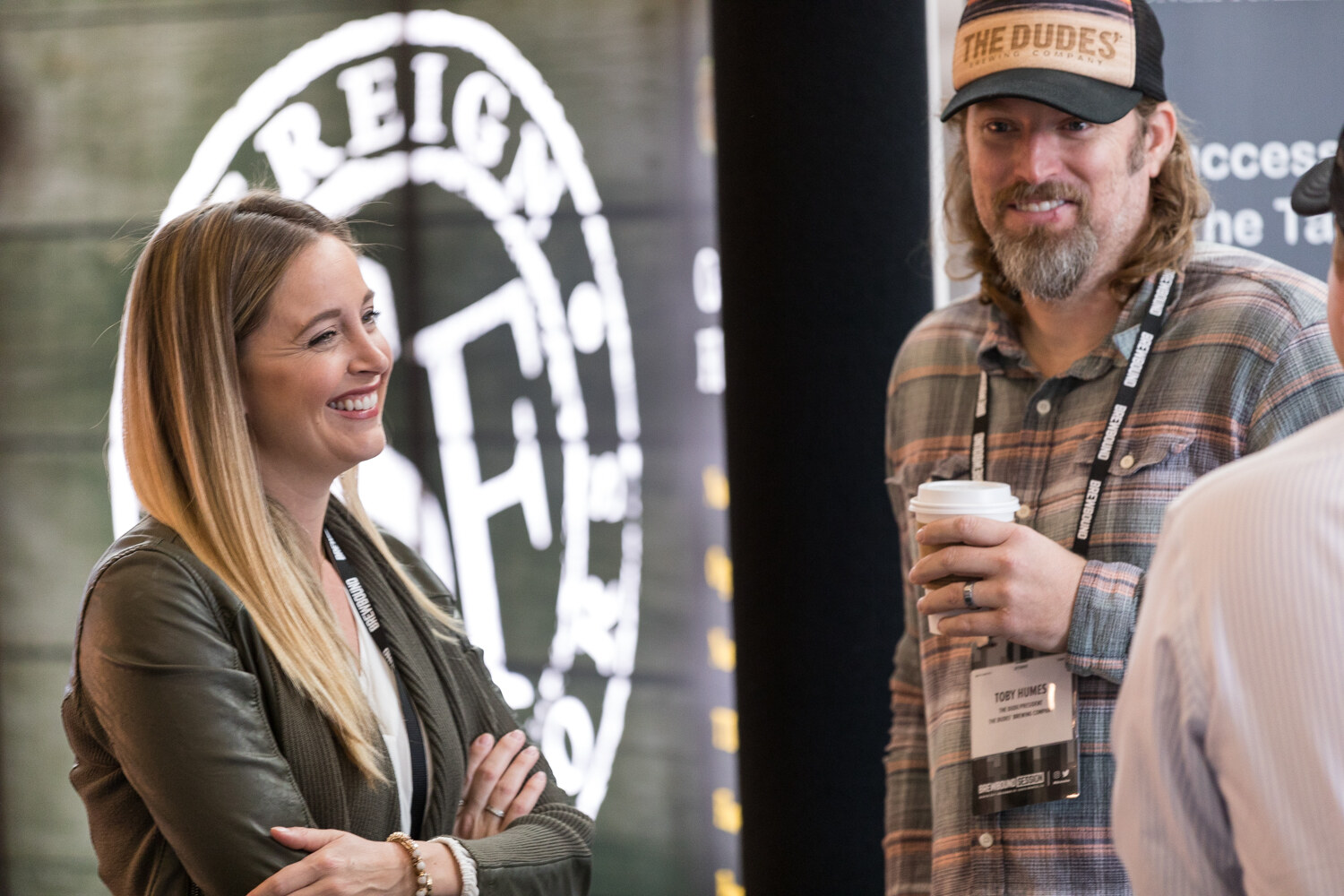 A smiling woman in an olive jacket with her arms crossed stands beside a bearded man in a plaid shirt and a trucker cap holding a coffee cup, with a large brewery logo banner behind them.