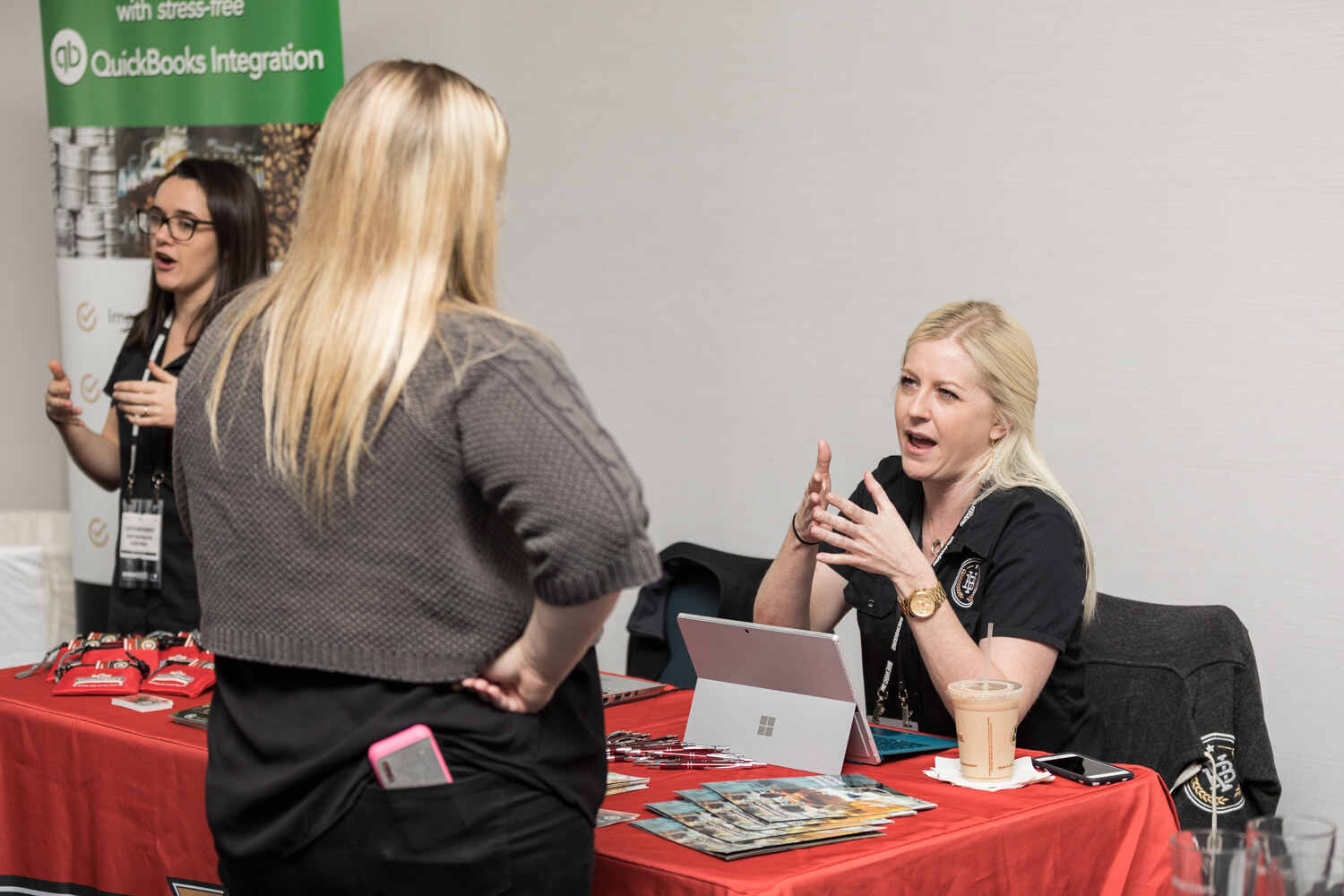 A woman in a black polo seated at a table with a red cloth, brochures and a Microsoft Surface laptop talks with a standing blonde visitor while a woman behind her stands in front of a QuickBooks Integration banner.
