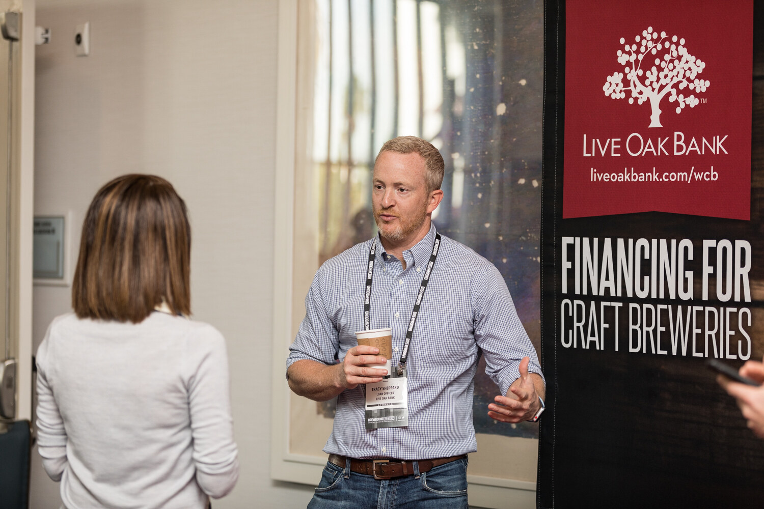 A man in a blue checked shirt holding a coffee cup gestures as he speaks to a woman with her back to the camera, beside a black banner reading Live Oak Bank Financing for Craft Breweries.
