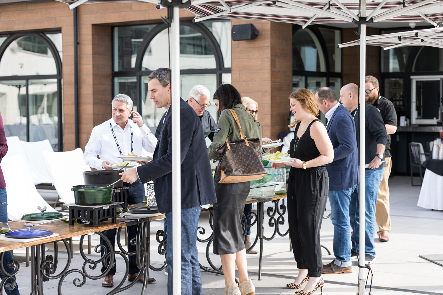 Attendees with lanyards line up with plates at a buffet of green cast-iron pots on an outdoor terrace under striped umbrellas.