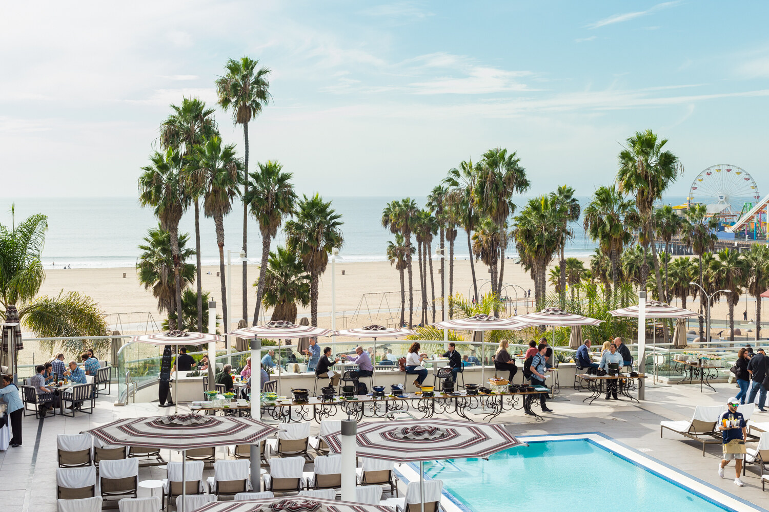 A wide view of a hotel pool deck with striped umbrellas, lounge chairs and a pool, palm trees, the beach and ocean, and a Ferris wheel on a pier at the right.