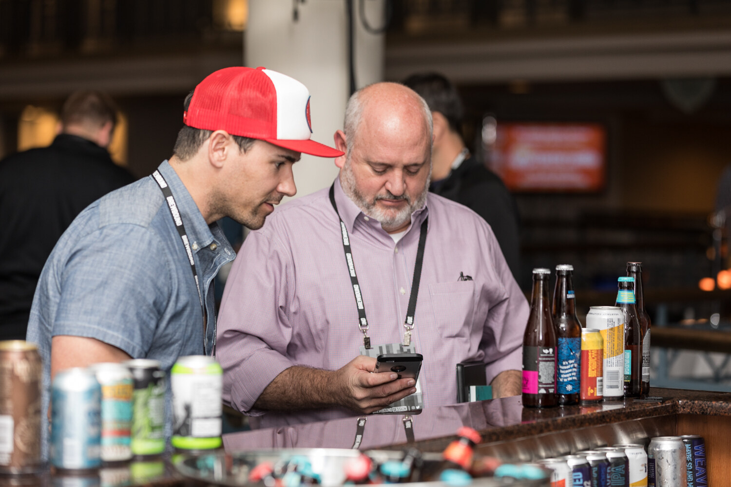 A man in a red and white trucker cap and a blue chambray shirt and a bearded man in a lilac shirt look at a phone over a granite bar lined with beer cans and bottles.