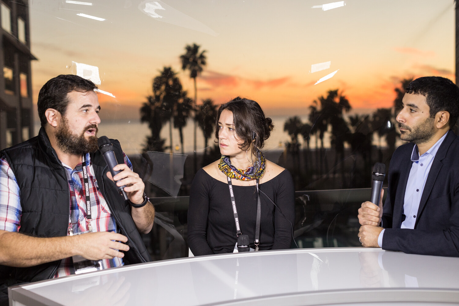 A bearded man in a black vest and plaid shirt speaks into a handheld microphone beside a woman in a black top with a colorful scarf and a man in a navy blazer holding a microphone, with silhouetted palm trees and an orange sky behind them.