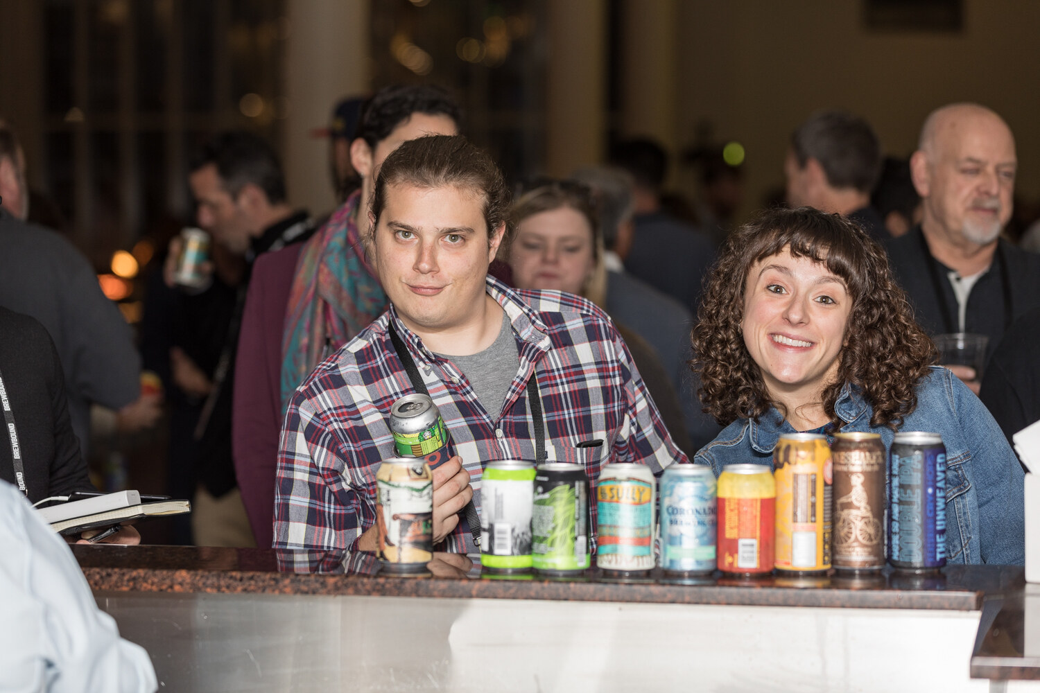 A man in a plaid shirt holding a can and a smiling woman in a denim jacket stand behind a bar with a row of beer cans lined up in front of them.