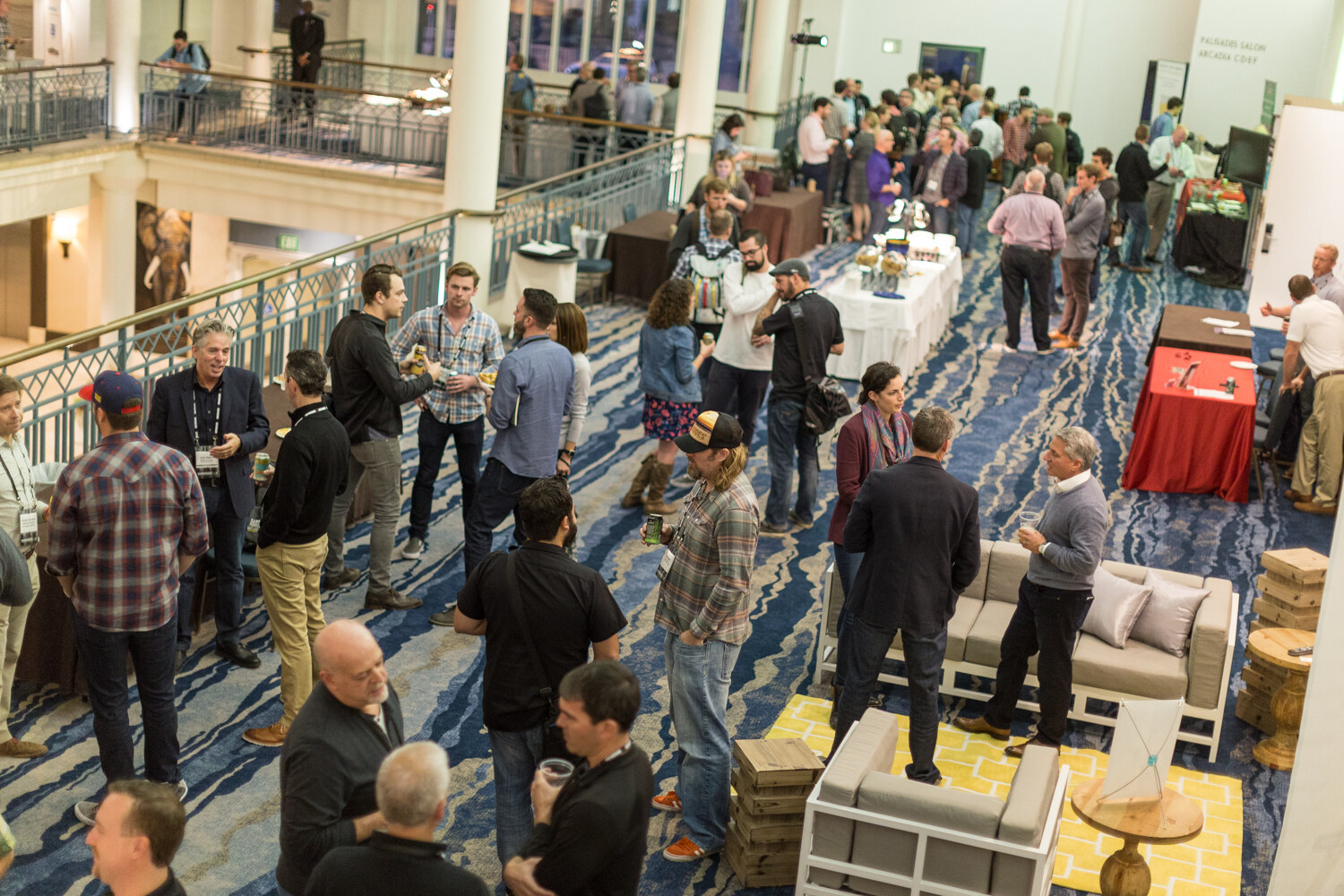 A high view of a hotel atrium where dozens of attendees talk in groups on a blue patterned carpet beside white outdoor-style sofas, a yellow rug and buffet tables.