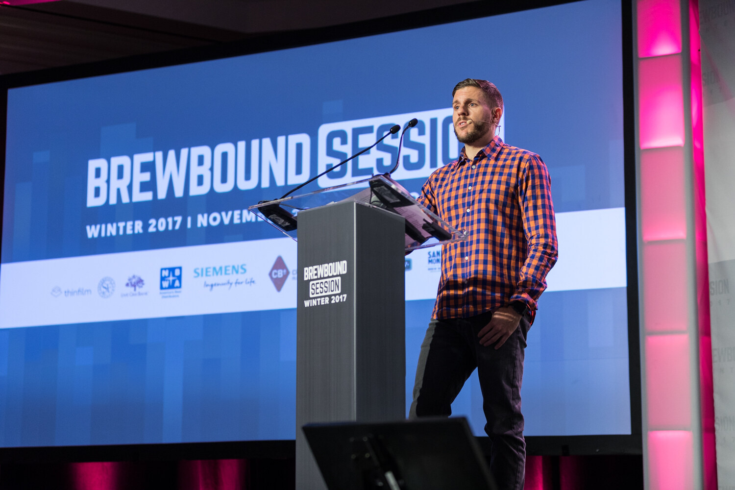 A bearded man in an orange and blue checked shirt stands beside a gray Brewbound Session Winter 2017 podium in front of a large blue screen with sponsor logos and magenta stage lighting.