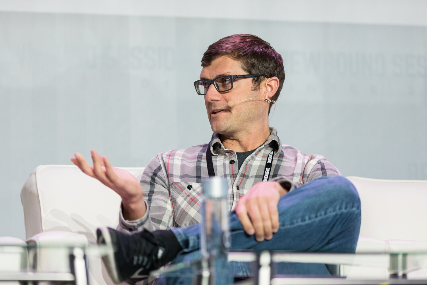 A man in black-framed glasses and a gray plaid shirt with a headset microphone sits with one leg crossed over the other and gestures with an open hand.