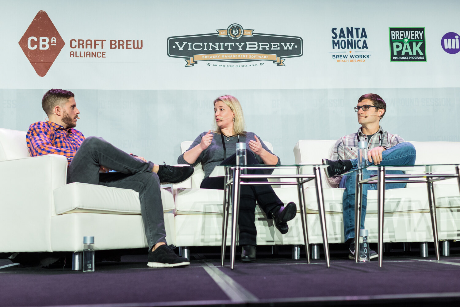 A host in an orange and blue checked shirt, a blonde woman in a gray sweater and a man in glasses and a plaid shirt sit on white sofas with glass tables in front of Craft Brew Alliance, Vicinity Brew, Santa Monica Brew Works and Brewery Pak logos.