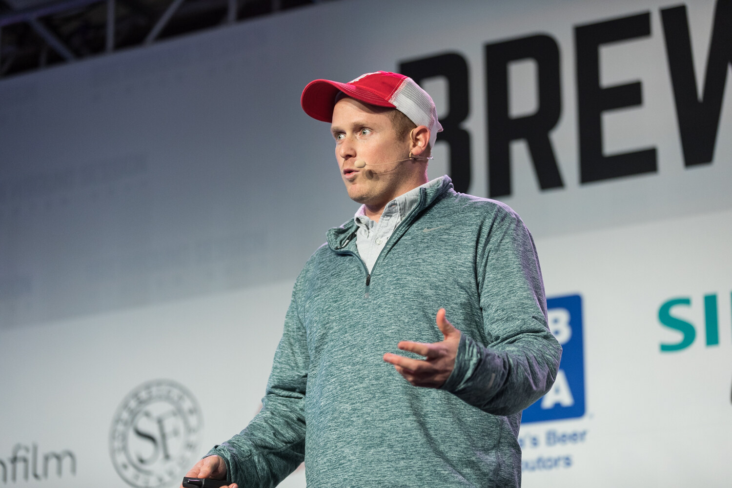 A man in a red and white trucker cap and a teal quarter-zip pullover with a headset microphone gestures in front of a Brewbound Session backdrop with Siemens and thinfilm logos.