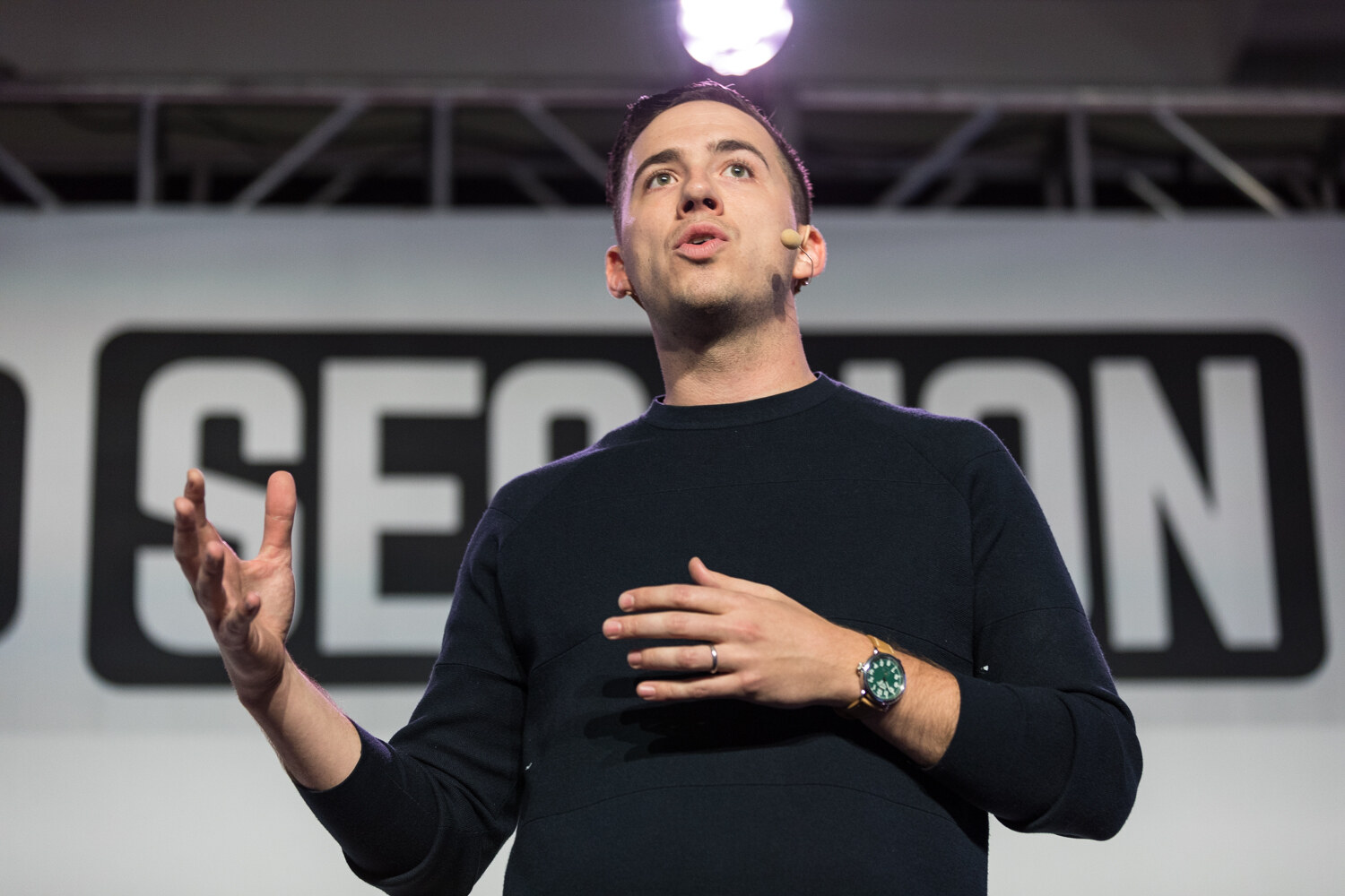 A man in a dark navy sweater and green-faced watch with a headset microphone gestures with both hands in front of the words Session on the backdrop.
