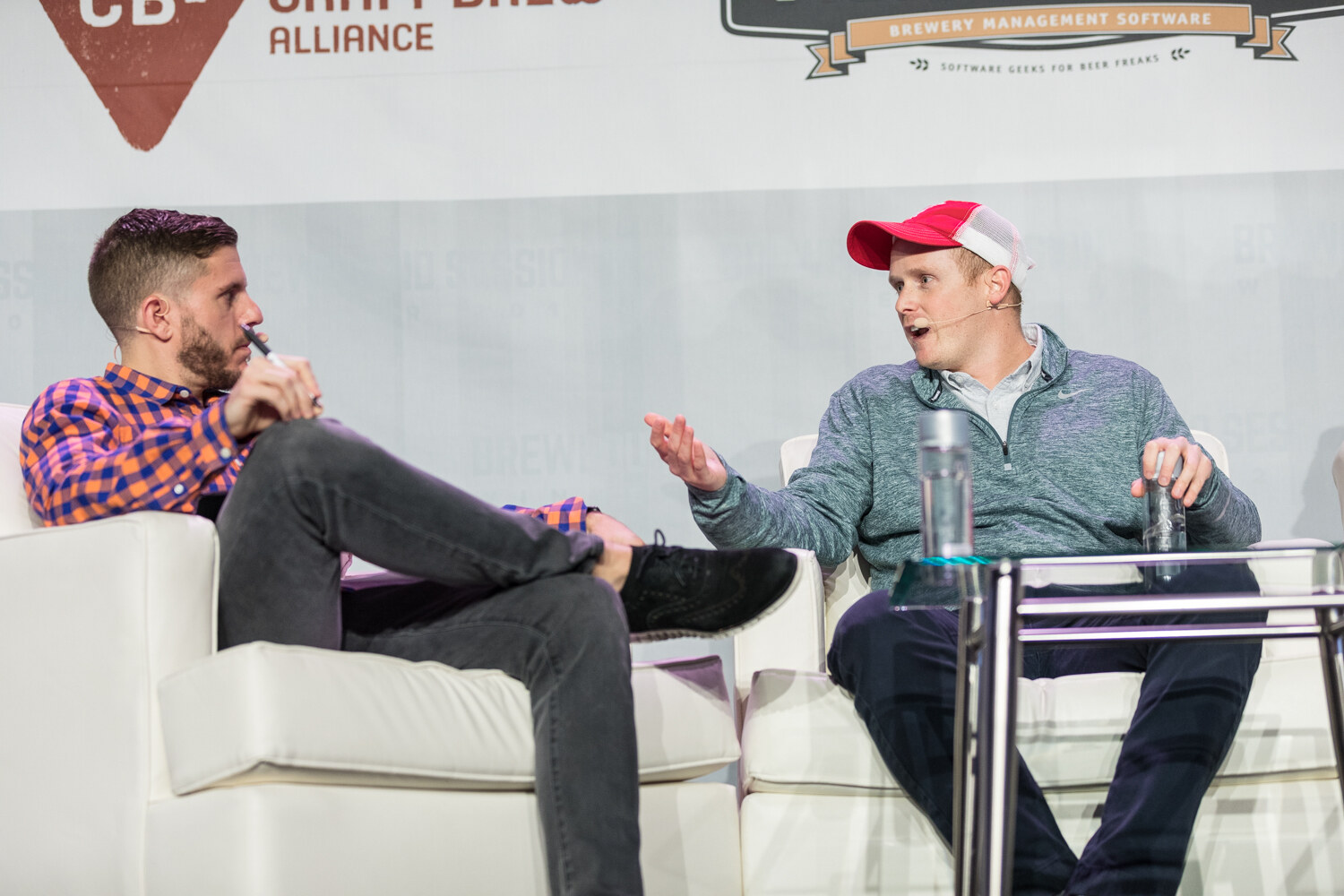 A host in an orange and blue checked shirt holding a pen sits on a white sofa while a man in a red trucker cap and teal pullover gestures toward him, in front of a Craft Brew Alliance banner.