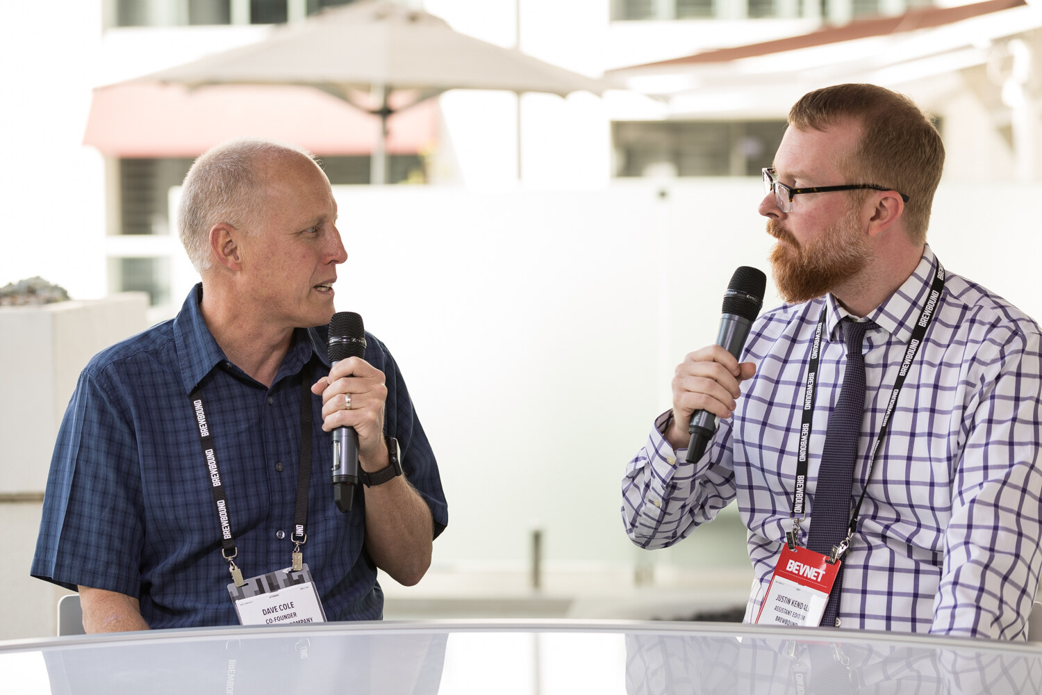 A man in a blue plaid shirt and a bearded man in a purple checked shirt with a BevNET lanyard hold microphones at a white curved desk, with a patio umbrella and hotel building behind them.