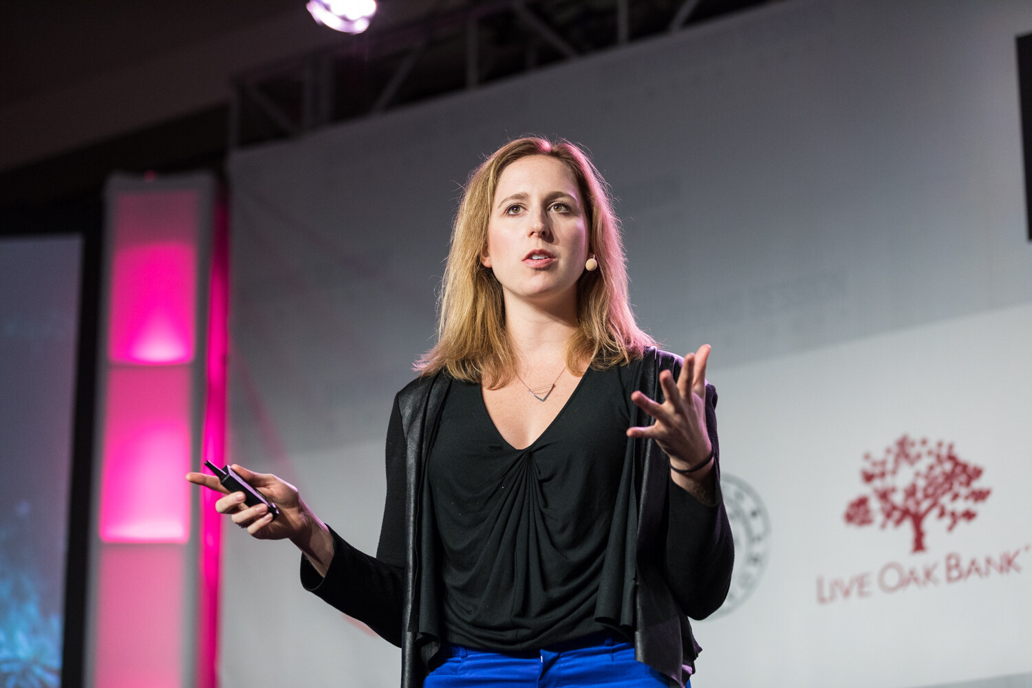A woman with shoulder-length blonde hair in a black cardigan and blue pants holds a clicker and gestures on stage in front of a Live Oak Bank logo and magenta stage lighting.