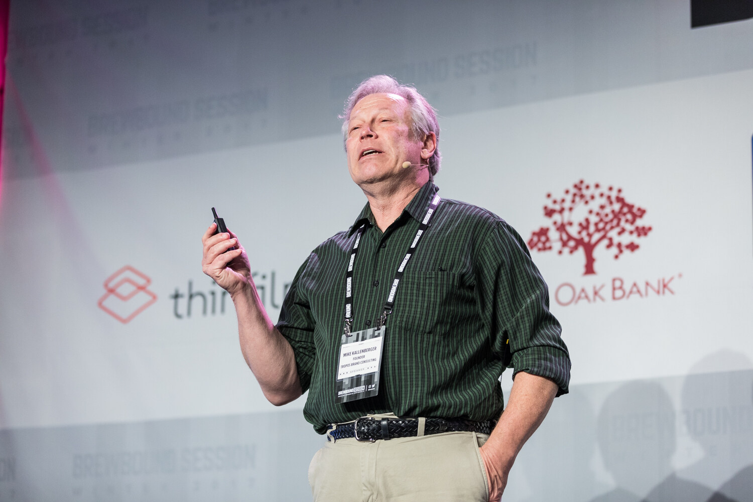 A gray-haired man in a green striped shirt and khakis with a headset microphone holds a clicker in front of thinfilm and Live Oak Bank logos.