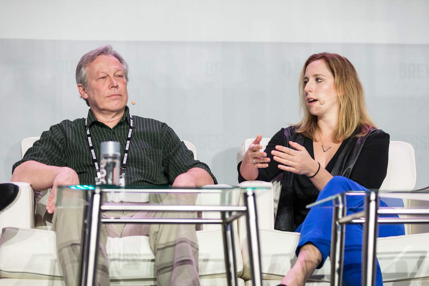 A man in a green striped shirt sits back in a white chair while a woman in a black top and blue pants gestures with both hands beside him.