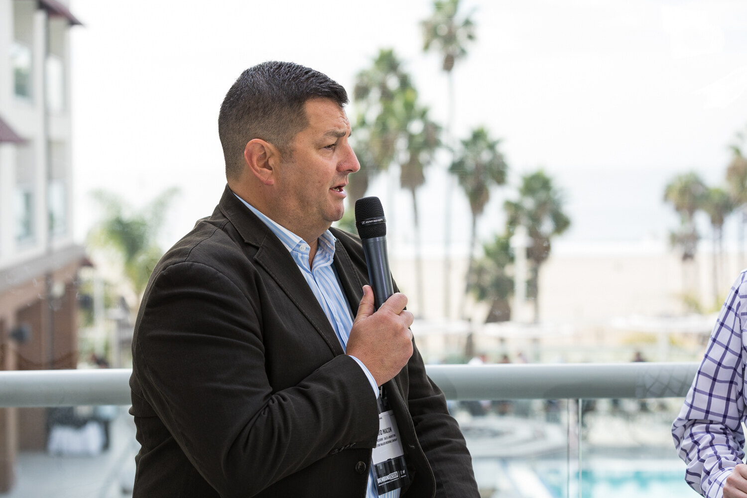 A man in a dark brown jacket and light blue striped shirt holds a microphone and speaks, with palm trees, a railing and the beach out of focus behind him.
