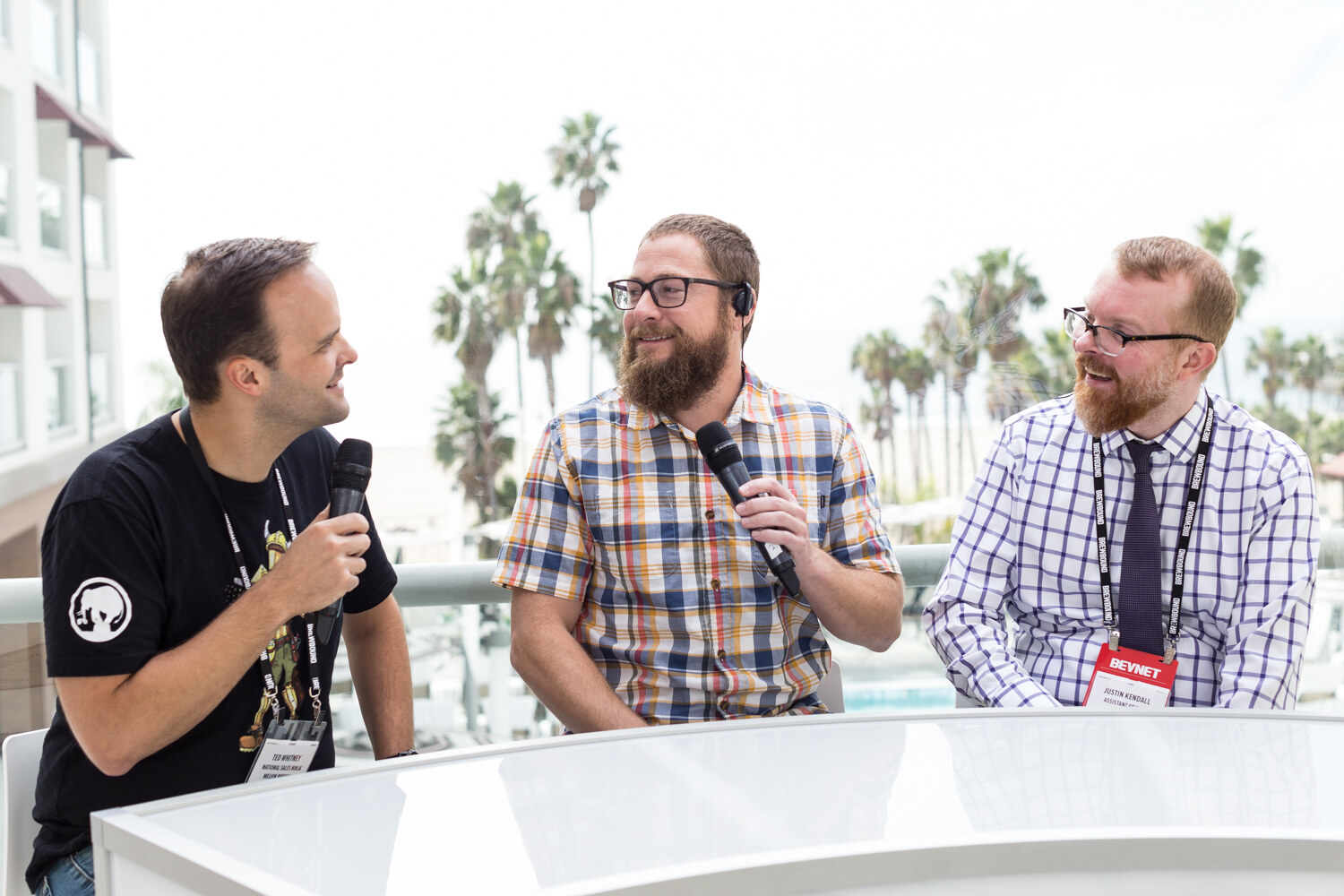 A man in a black T-shirt with a microphone, a bearded man in a plaid shirt with a headset and a man in a checked shirt with a BevNET lanyard sit at a white curved desk with palm trees and a pool behind them.