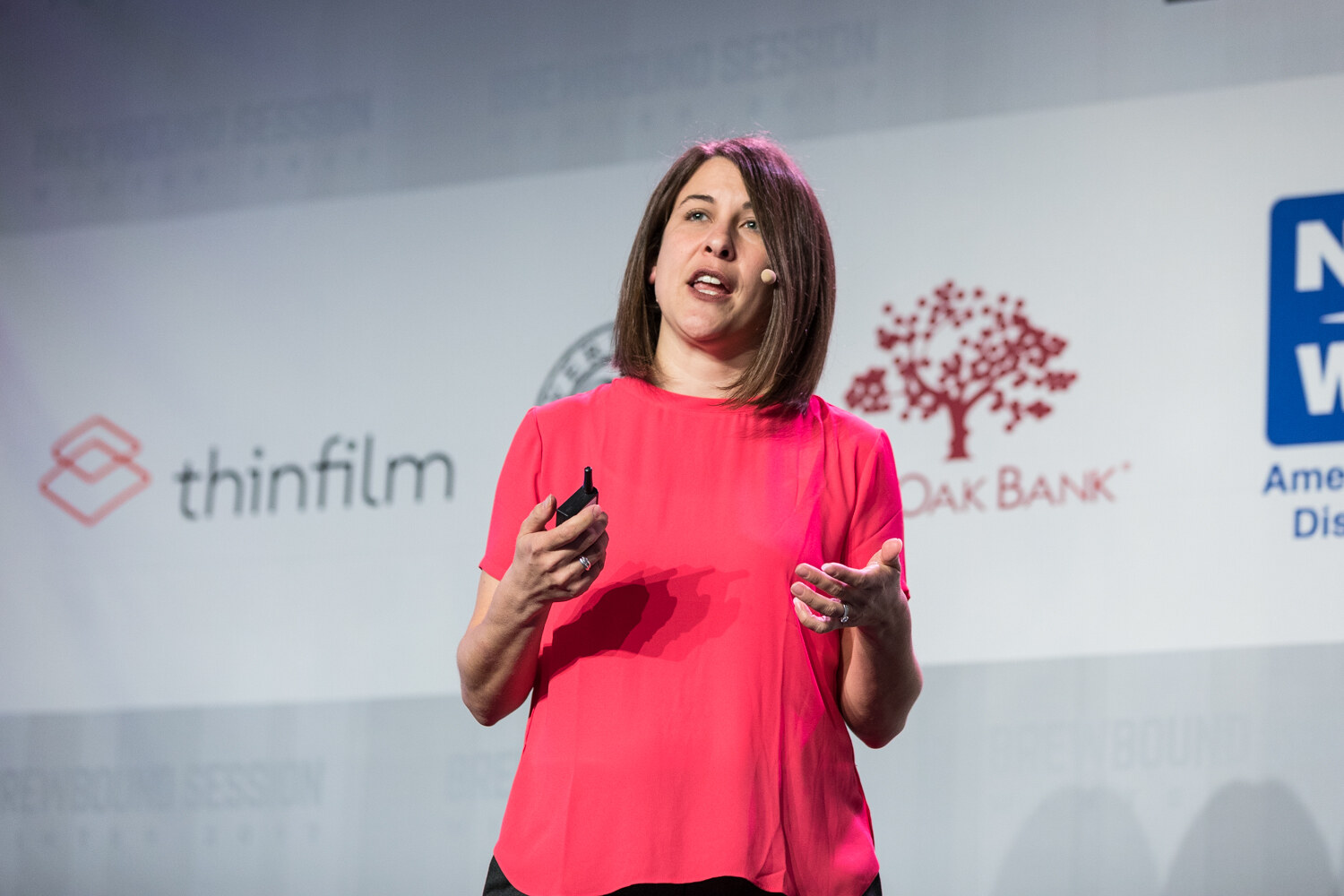 A woman with a brown bob in a coral top holds a clicker and speaks in front of thinfilm, Live Oak Bank and NBWA logos on the stage backdrop.