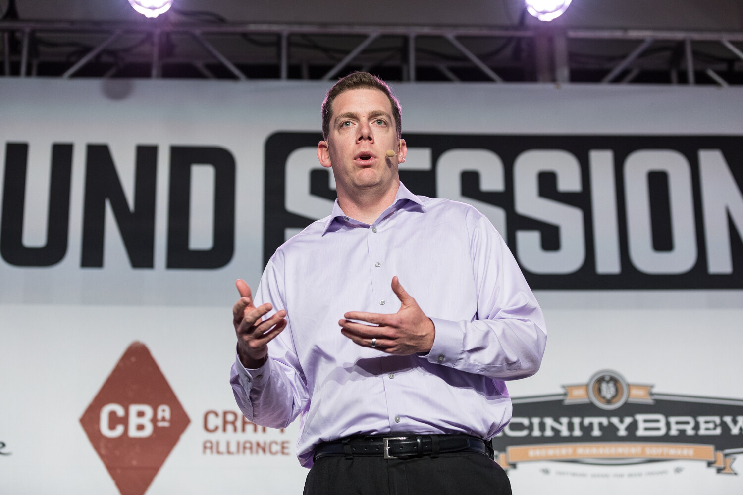 A man in a lavender button-up shirt and black belt with a headset microphone gestures with both hands in front of the Brewbound Session backdrop with Craft Brew Alliance and Vicinity Brew logos.