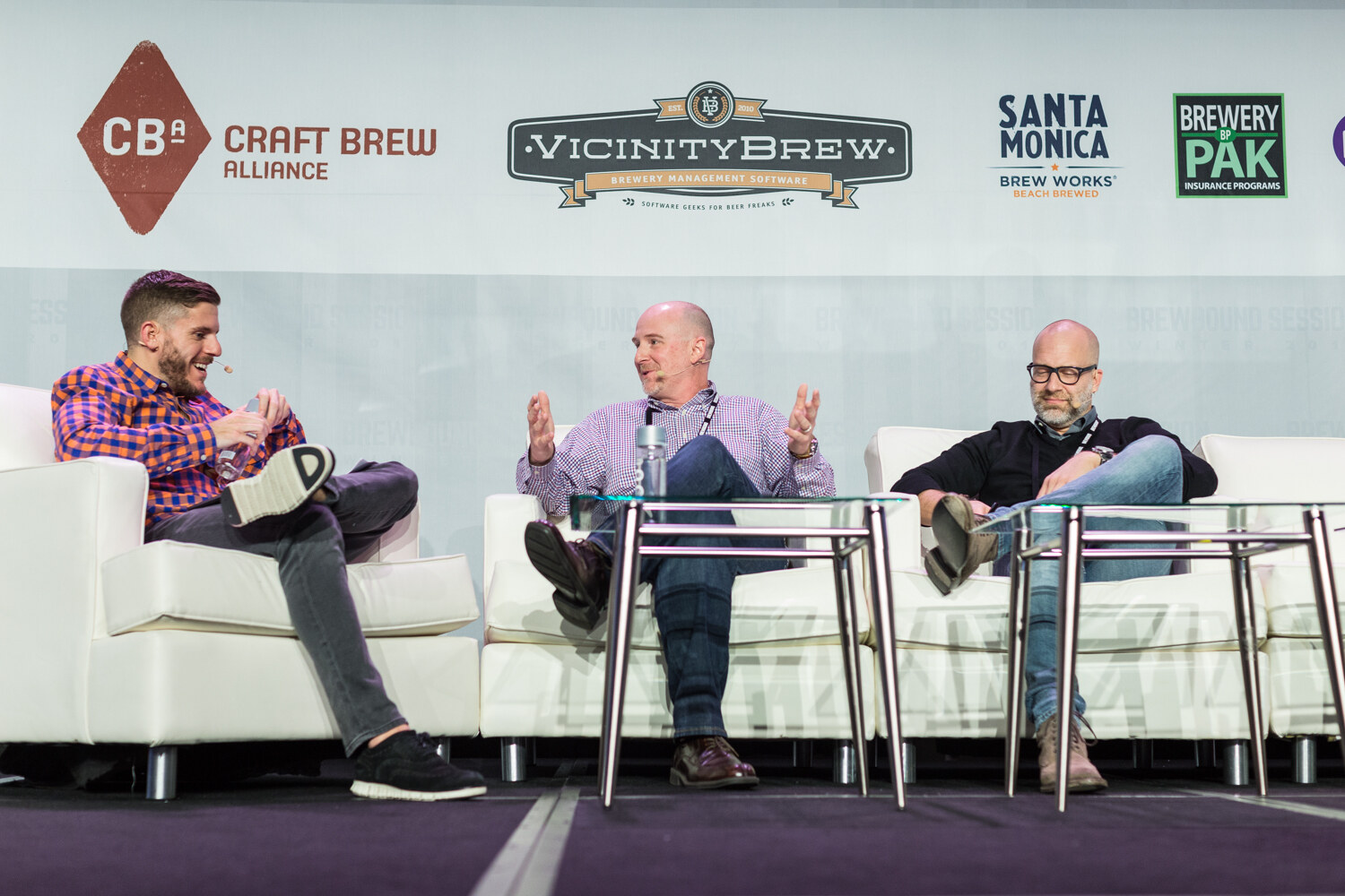 A host in an orange and blue checked shirt, a bald man in a purple checked shirt and a bald bearded man in a black sweater sit on white sofas in front of Craft Brew Alliance, Vicinity Brew, Santa Monica Brew Works and Brewery Pak logos.
