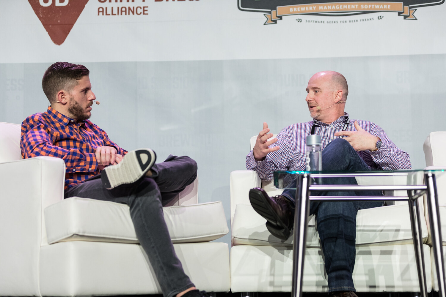 A host in an orange and blue checked shirt sits on a white sofa facing a bald man in a purple checked shirt who gestures with both hands, in front of a Craft Brew Alliance banner.