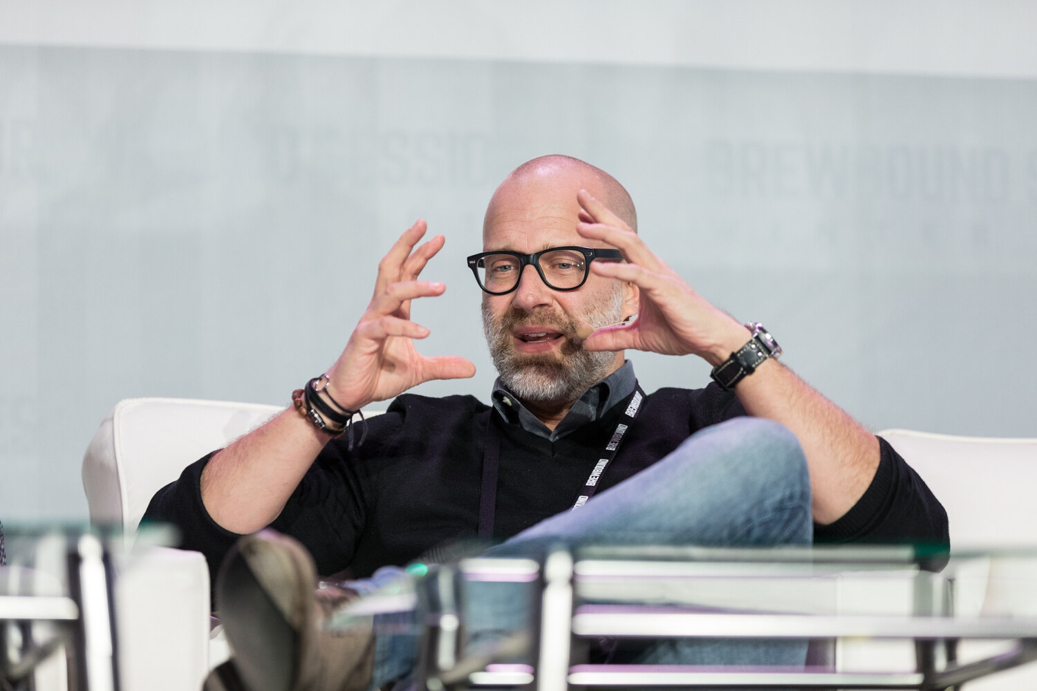 A bald bearded man in black-framed glasses and a black sweater holds both hands near his face while speaking, seated on a white sofa.