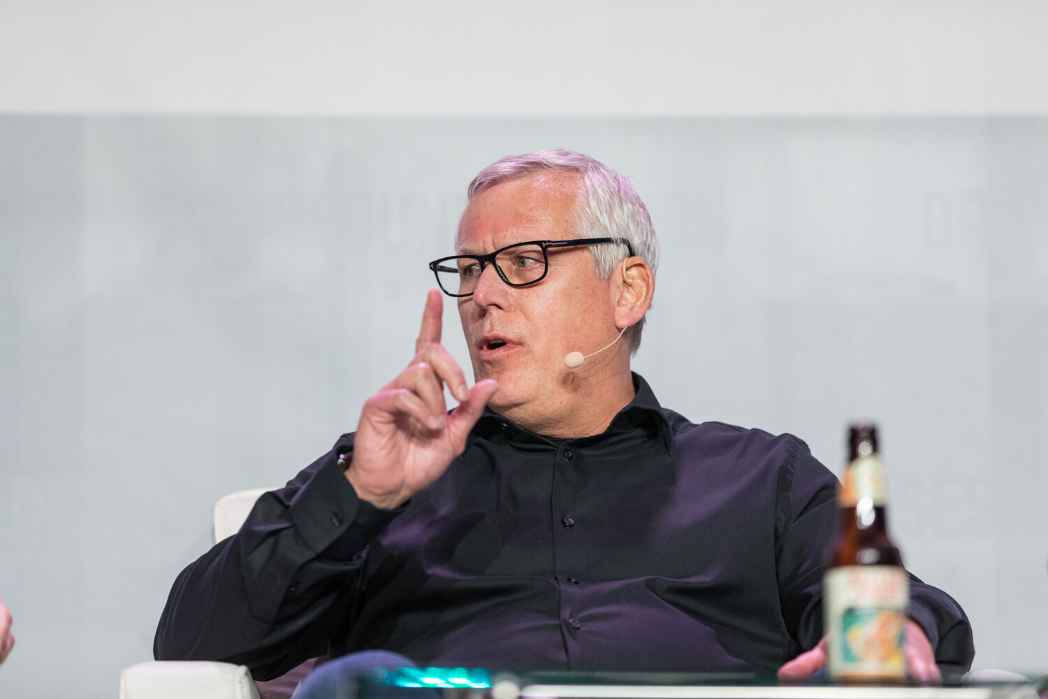 A gray-haired man in glasses and a black button-up shirt with a headset microphone raises one finger while speaking, with a beer bottle on the table in front of him.
