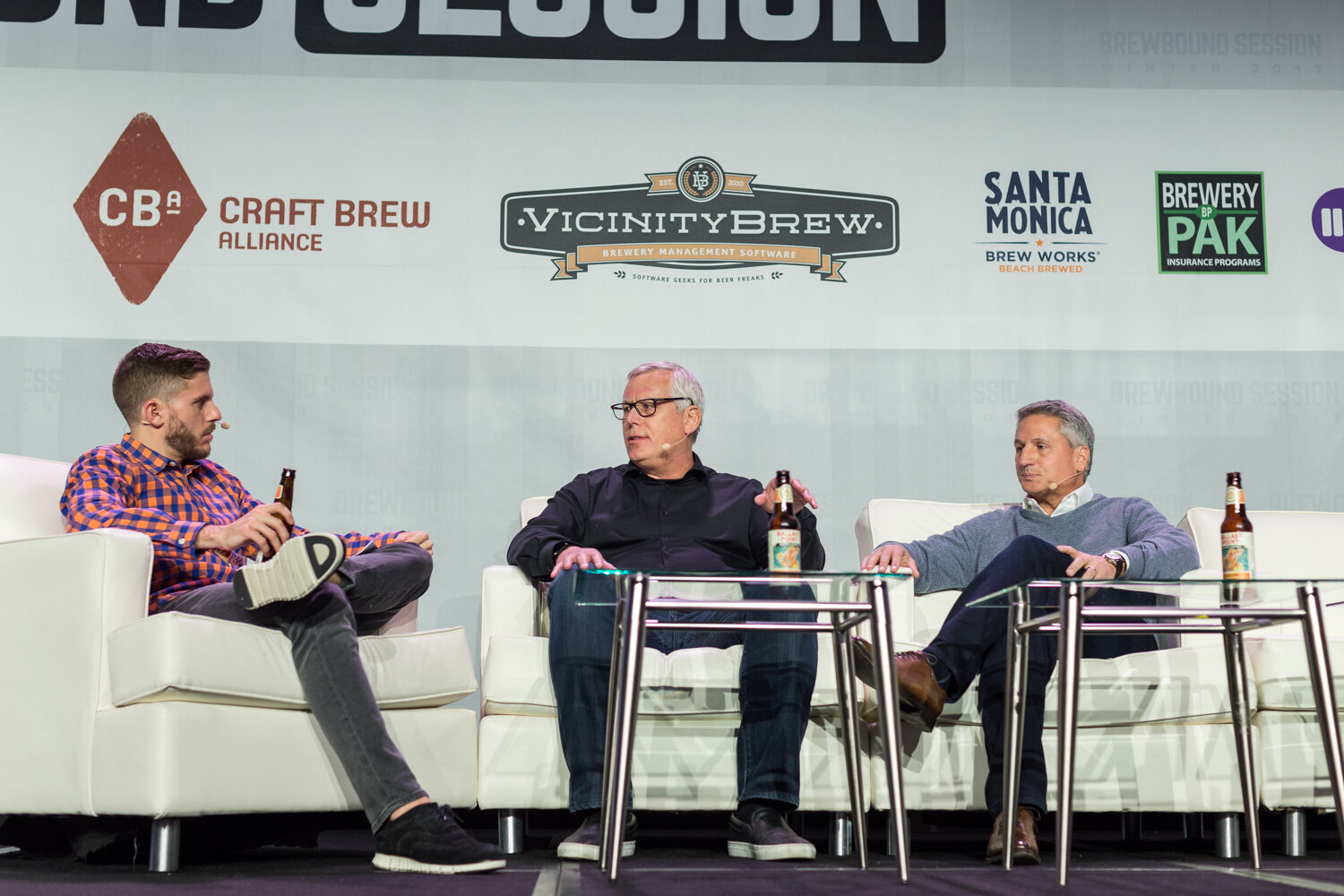 A host in an orange and blue checked shirt and two men, one in a black shirt and one in a gray sweater, sit on white sofas with beer bottles on glass tables in front of Craft Brew Alliance, Vicinity Brew, Santa Monica Brew Works and Brewery Pak logos.
