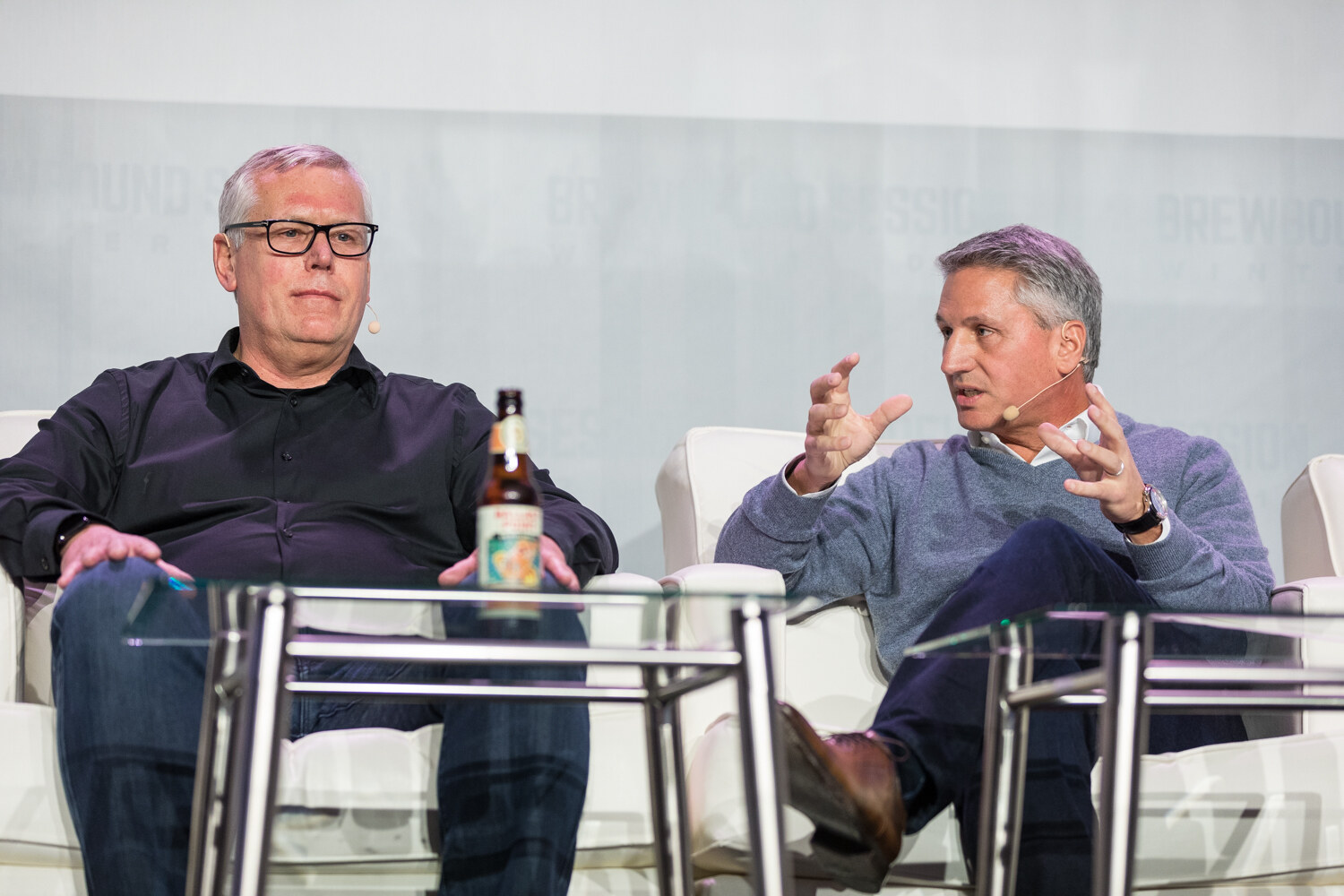 A man in a black shirt and glasses sits listening beside a man in a gray sweater who gestures with both hands, with a beer bottle on the glass table in front of them.