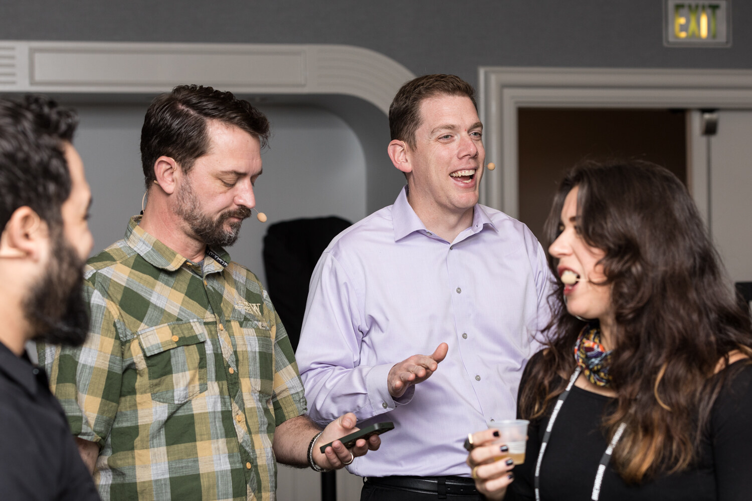 A man in a green plaid shirt looks down at a phone, a man in a lavender shirt laughs and gestures, and a dark-haired woman with a colorful scarf turns toward him, while a bearded man is partly visible at the left edge of the frame.
