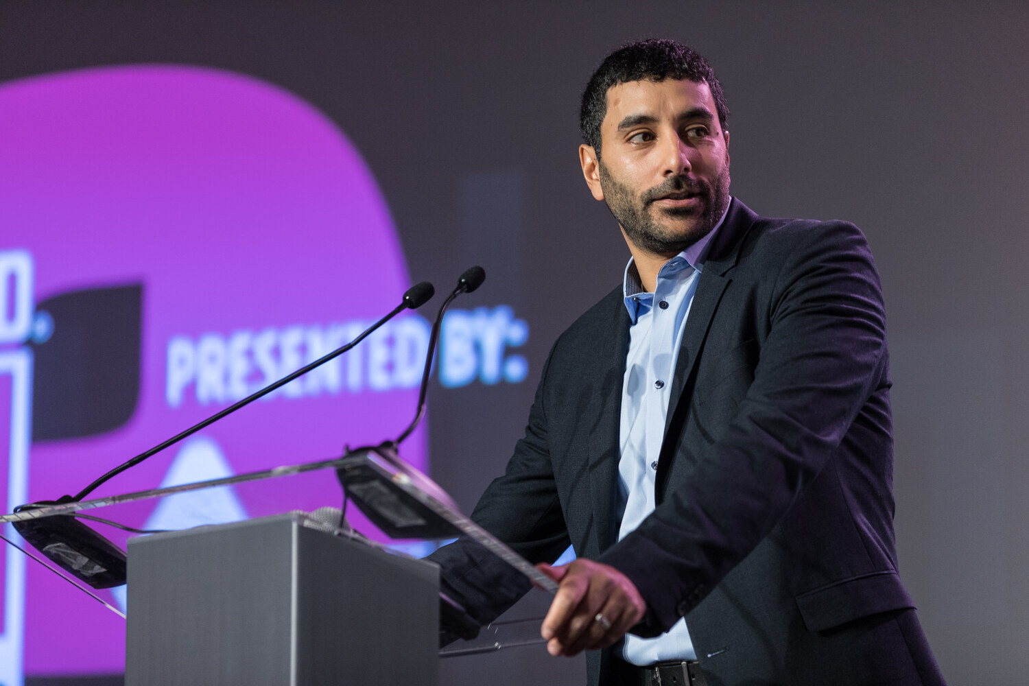 A man in a navy blazer and light blue shirt looks to his left at a clear podium with two microphones, in front of a purple Presented By slide.