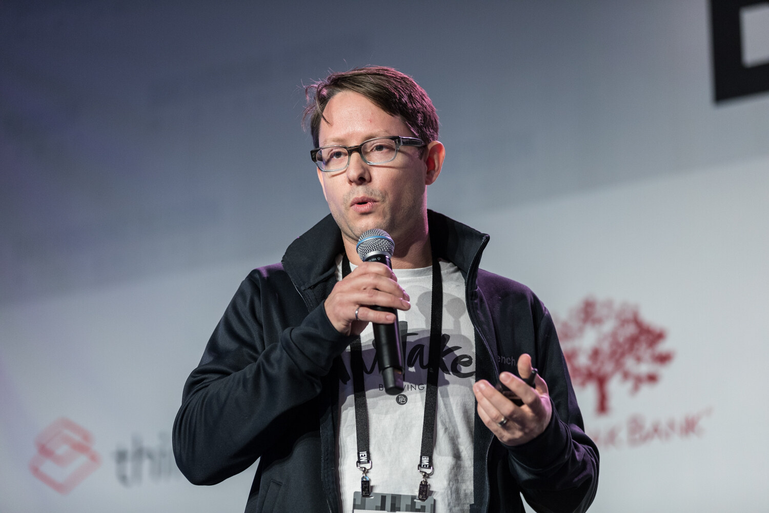 A man in glasses and a black zip jacket over a gray Brewing T-shirt holds a handheld microphone and a clicker in front of thinfilm and Live Oak Bank logos.