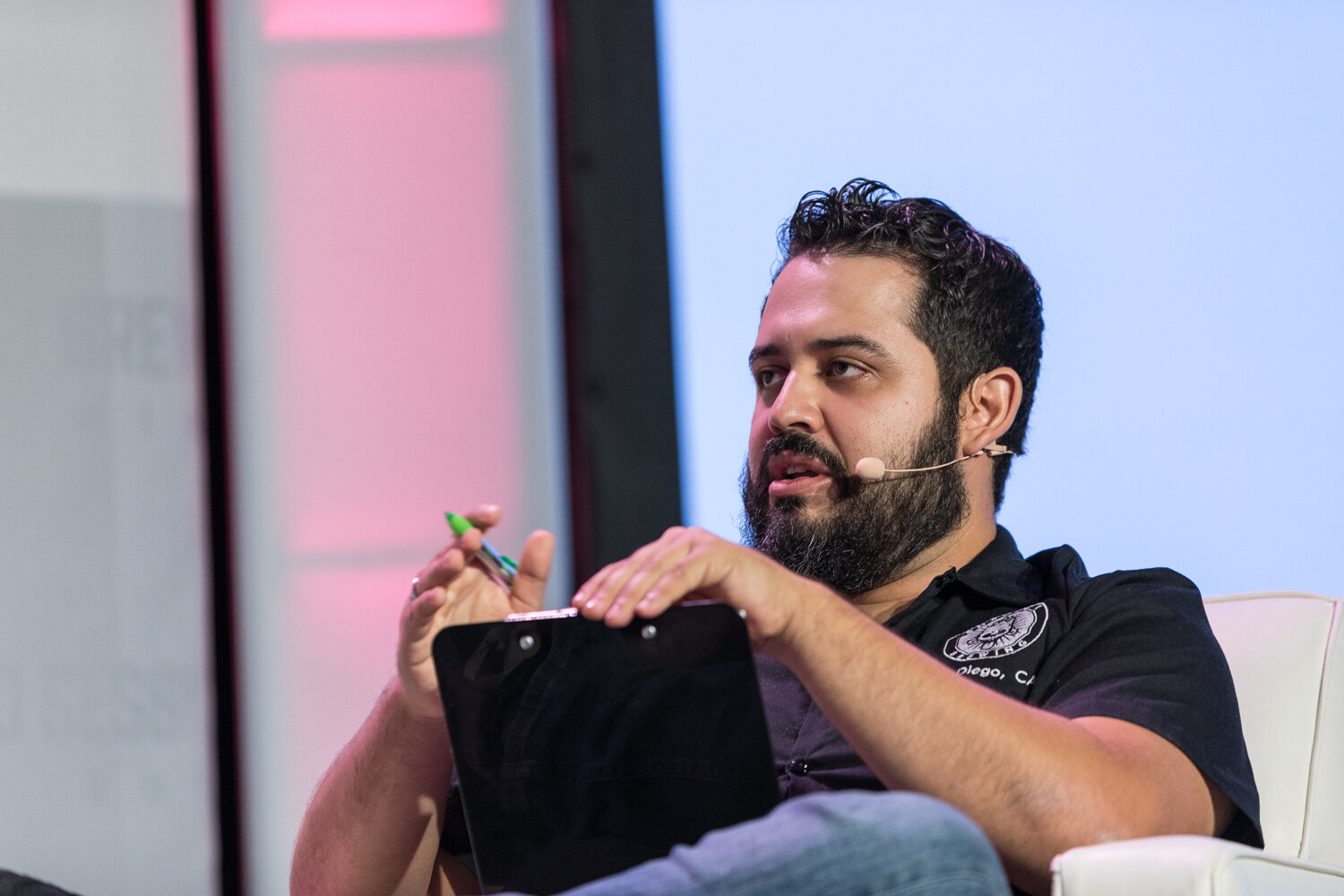 A bearded man in a black polo with a headset microphone holds a pen over a clipboard while seated on a white chair in front of pink and blue stage lighting.
